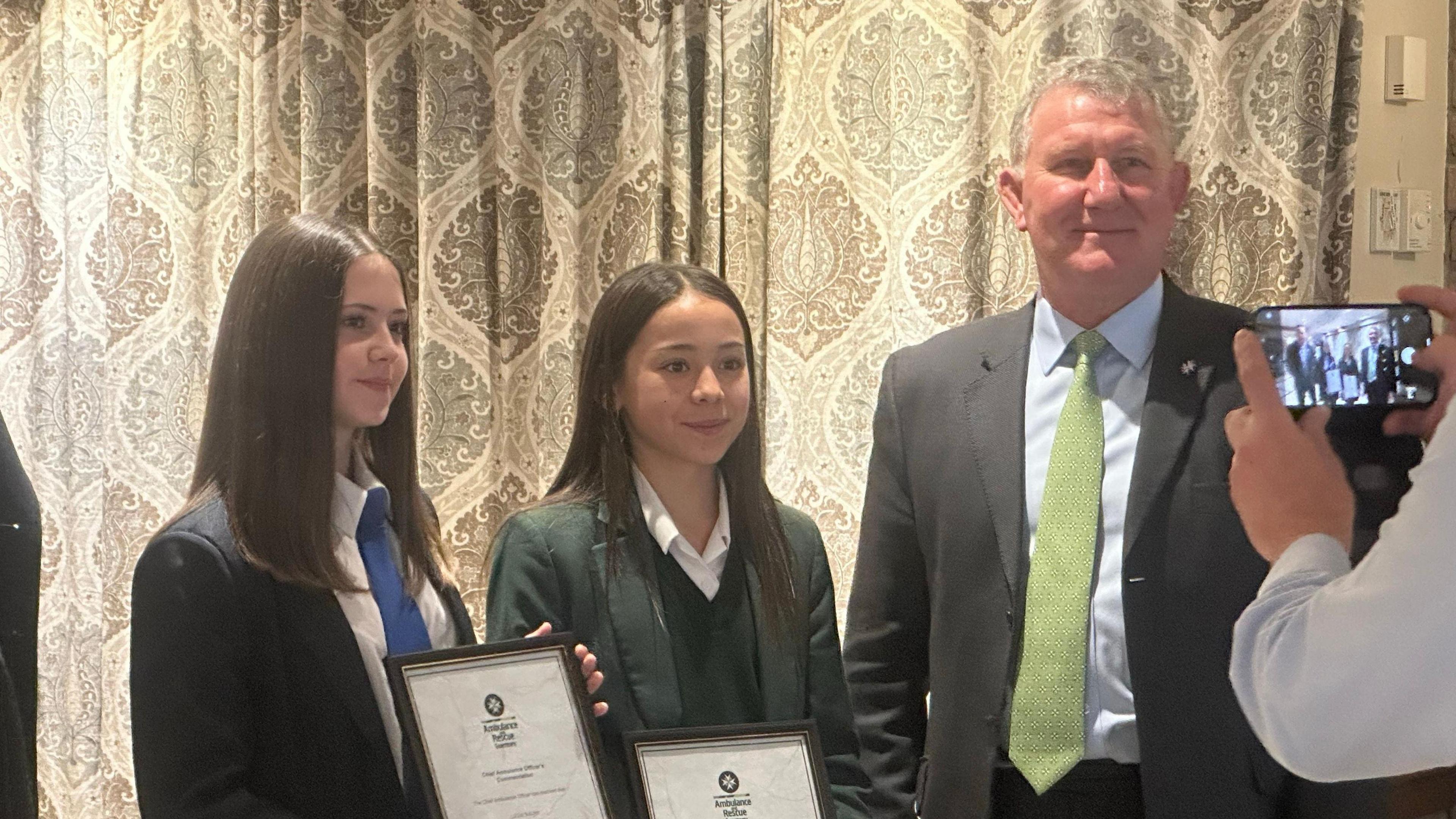 Juliette and Lillia stood in school uniforms with the framed awards in their hands. There is a man stood to the right and they are all posing for a photograph. 