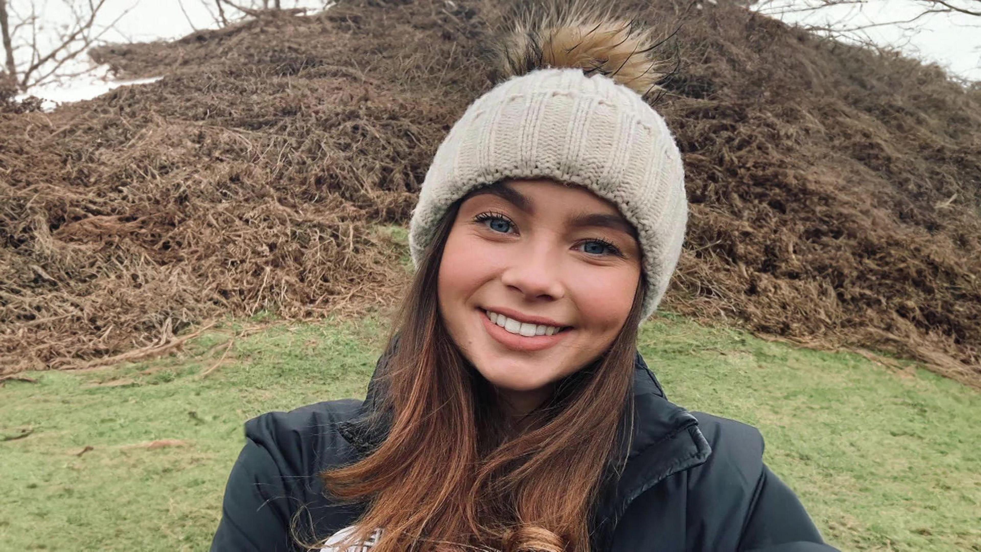 Jaysley smiling at the camera. She has long dark hair and is wearing a black coat and a cream woolly hat with a brown pompom on top. 