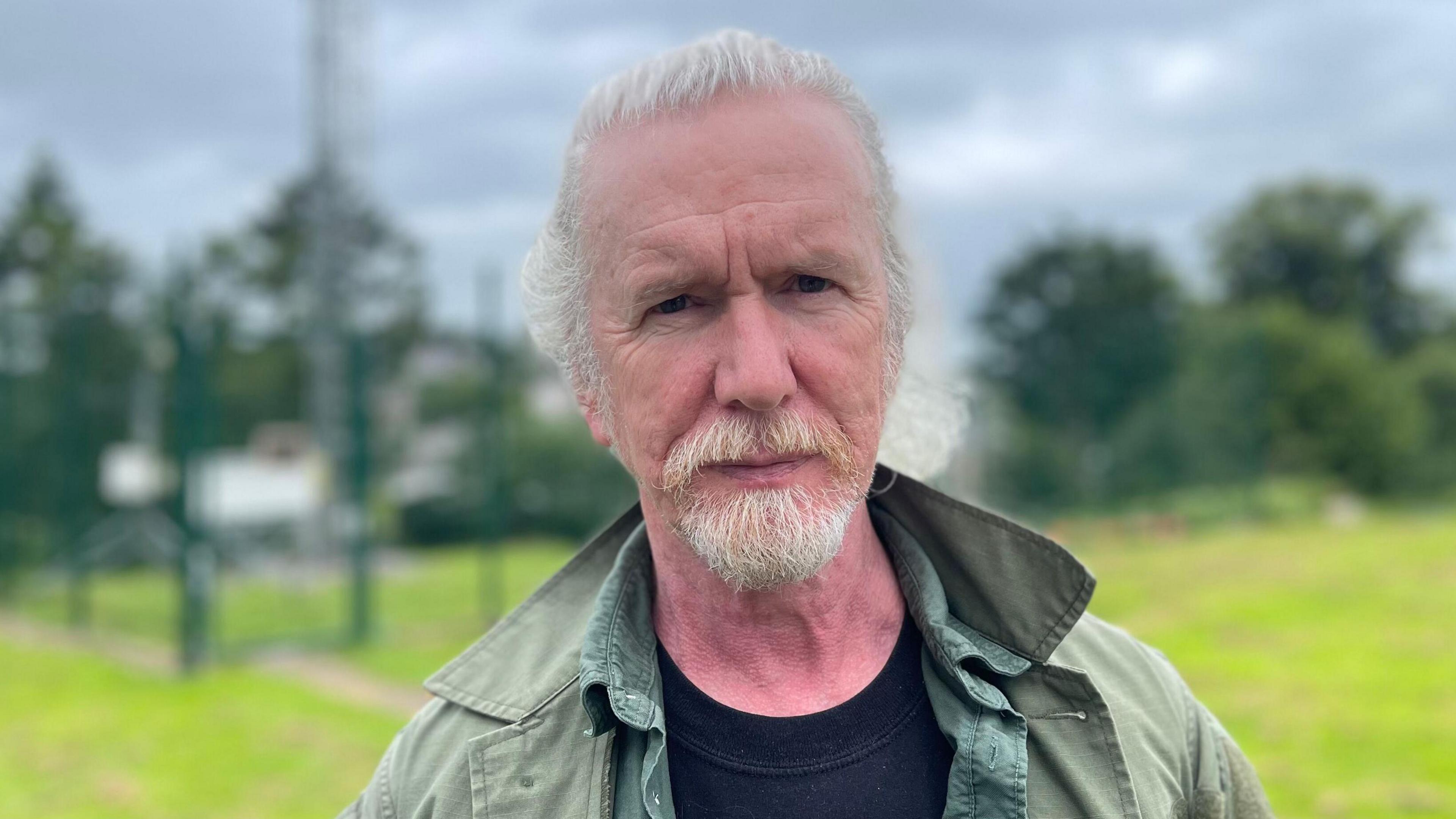 Shane Kelly, a man with white hair and beard, looks into the camera. He is wearing a green coat over a black t-shirt and a green shirt.