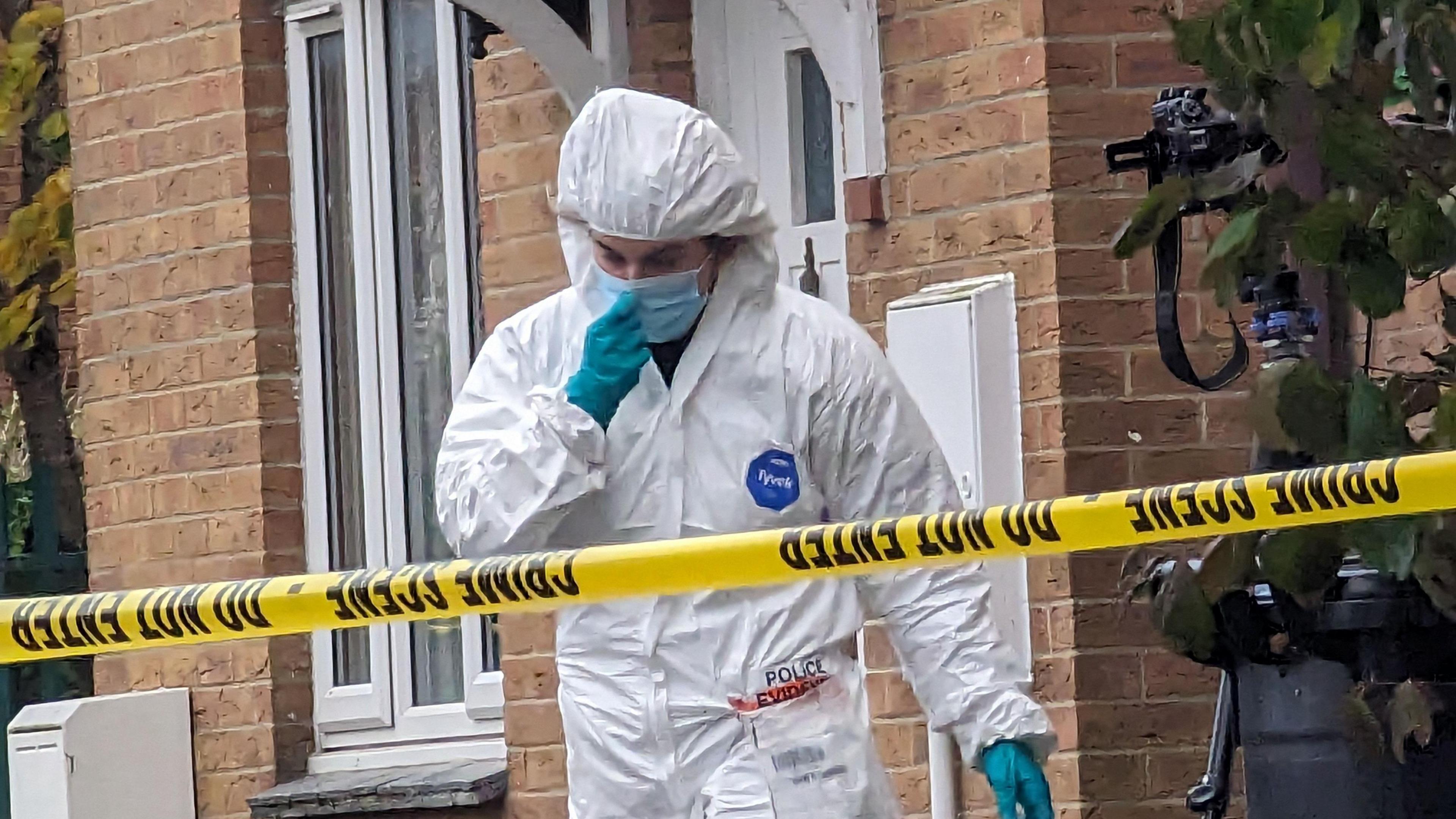 A forensics officer in a white boiler suit walks out of a house