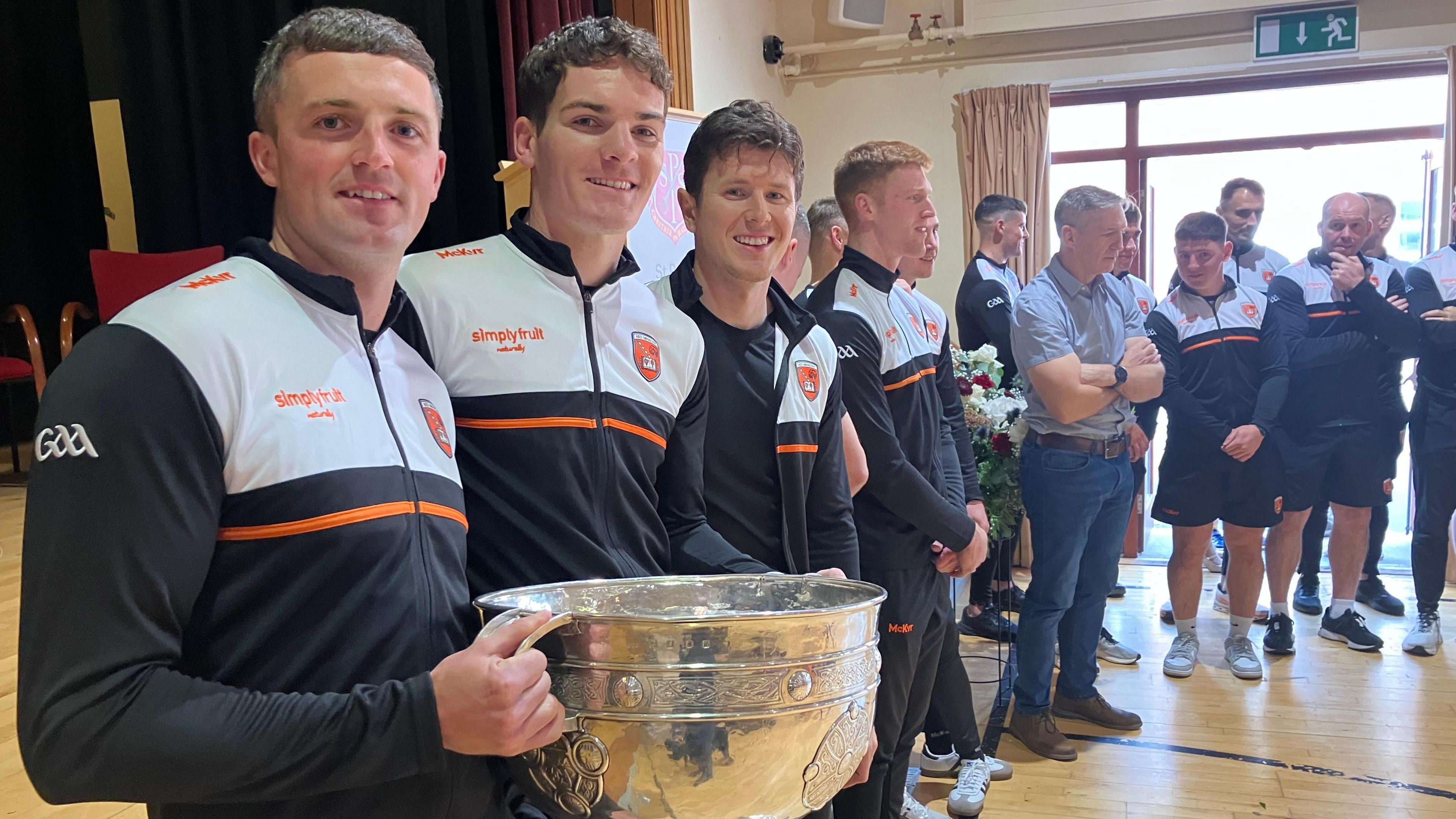 Group of Armagh players in their team hit holding the Sam Maguire cup in a school hall