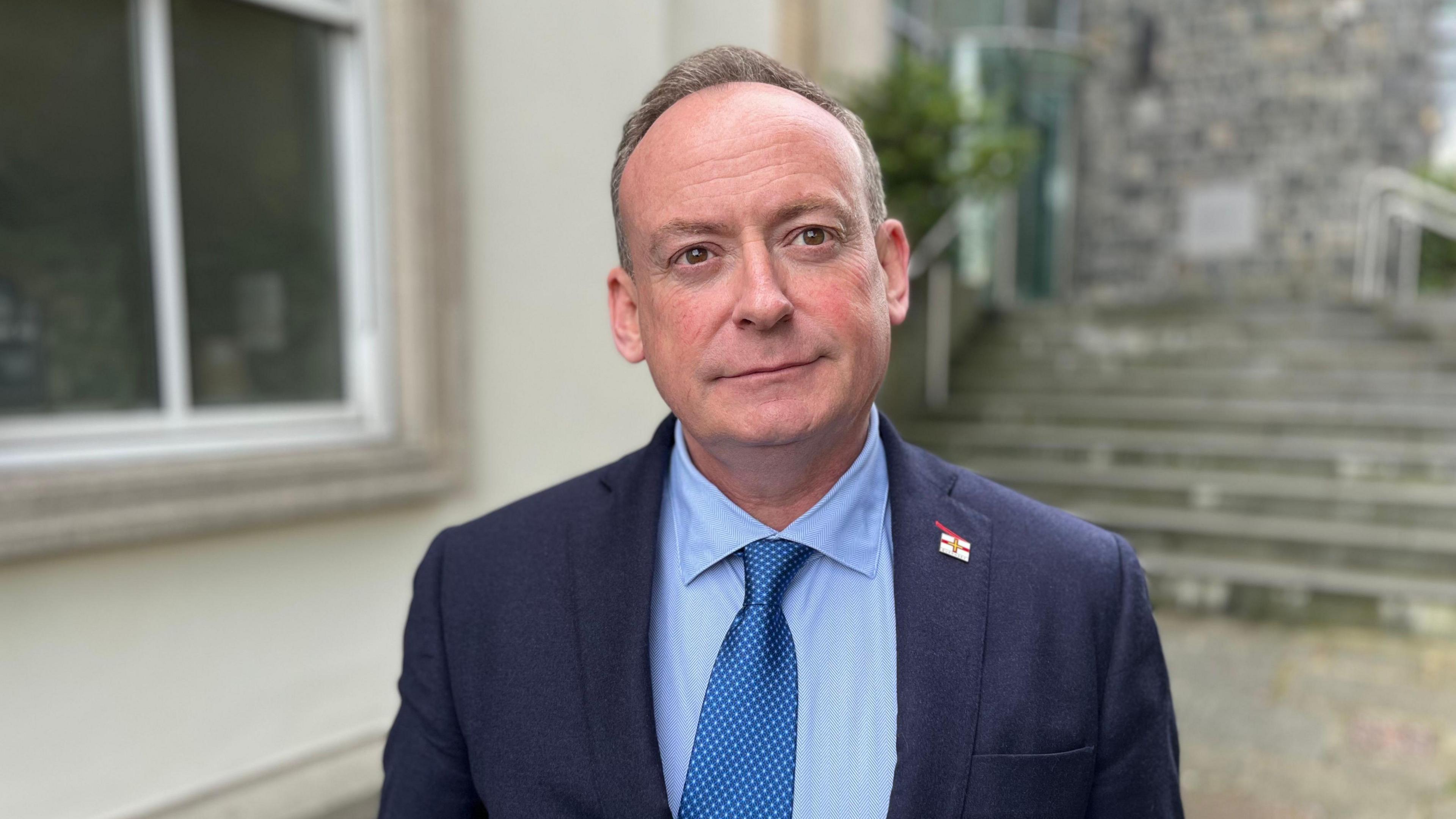 A man wearing a blue shirt and a blue suit with a metallic blue tie looking towards the camera. On his left lapel he is wearing a Guernsey flag pin badge. 