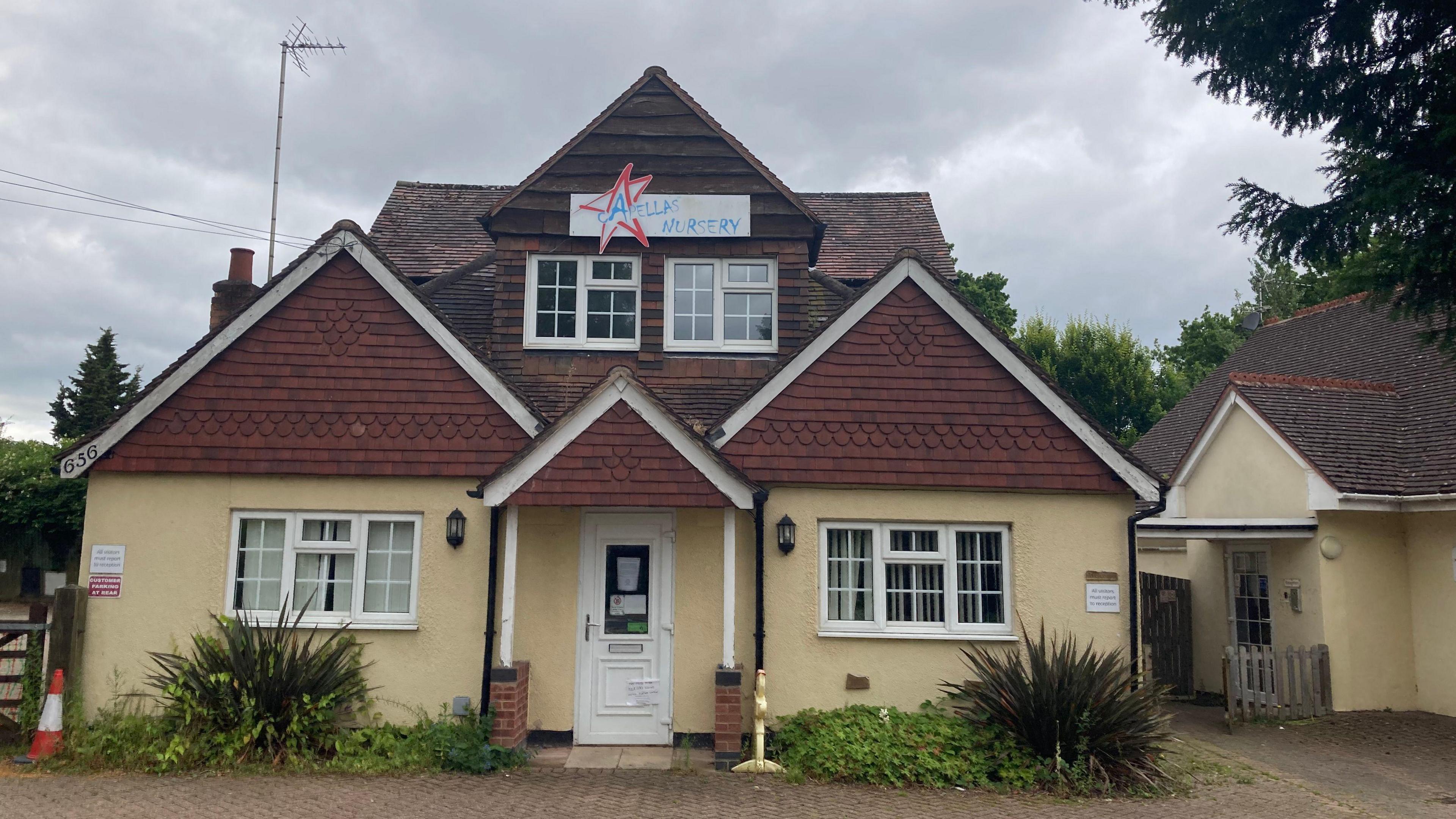 The front of a nursery building. The walls are yellow/beige, with brown brick on pointed parts of the roof. There is a sign in the centre of the roof reading "Capellas nursery"