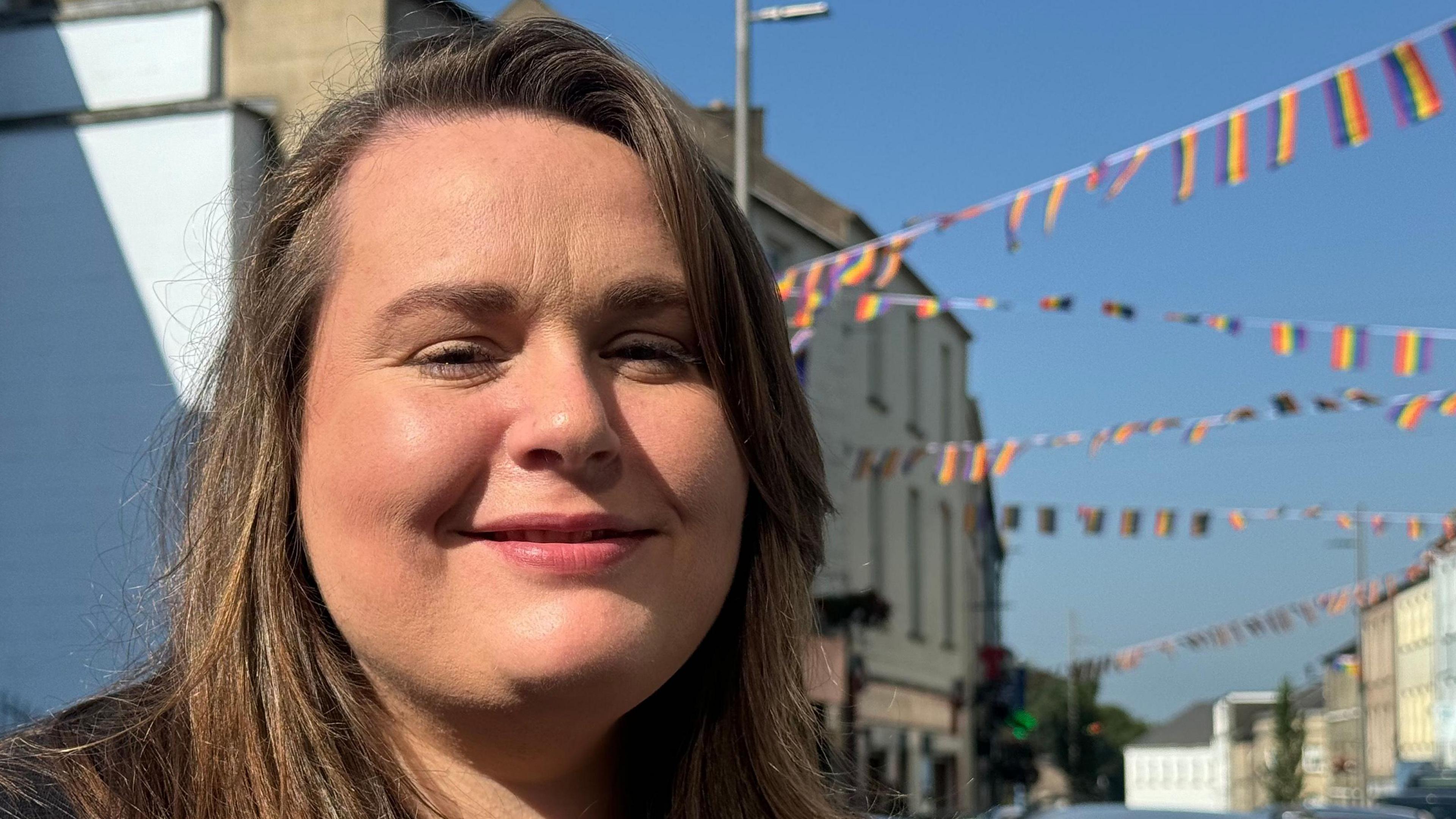 Bernie has brown hair. Behind her is bunting with rainbow colours. It's hanging from one end of the street to the other.