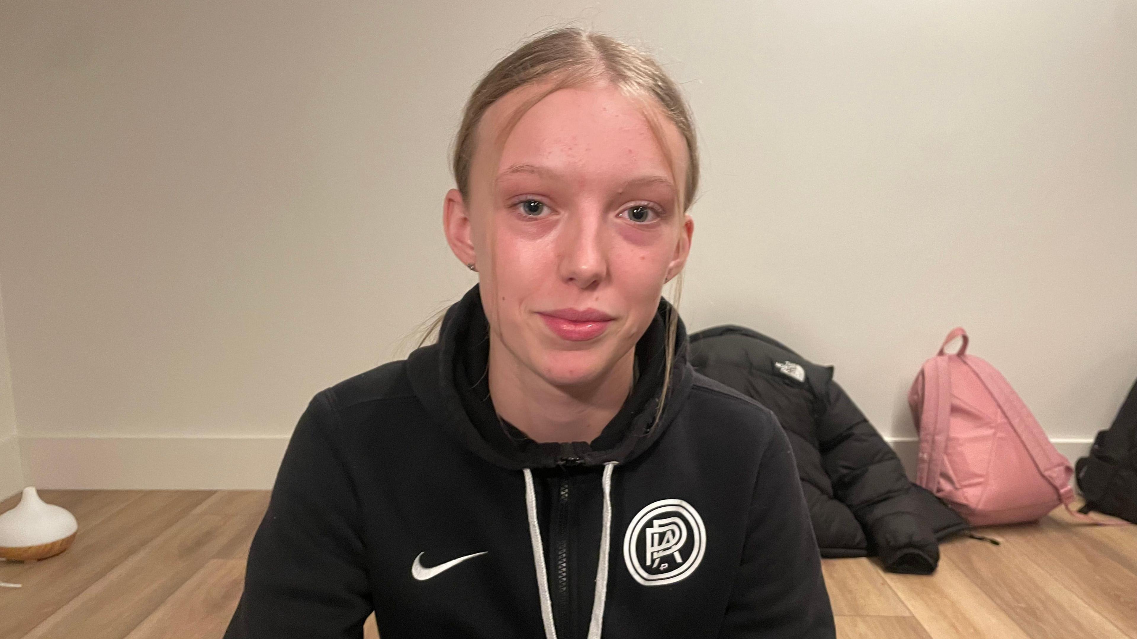 16-year-old Annie, is smiling towards the camera and wears a nike black zip up hoodie. Annie has blonde hair pulled back with two face framing stands of hair. Annie has blue eyes. 