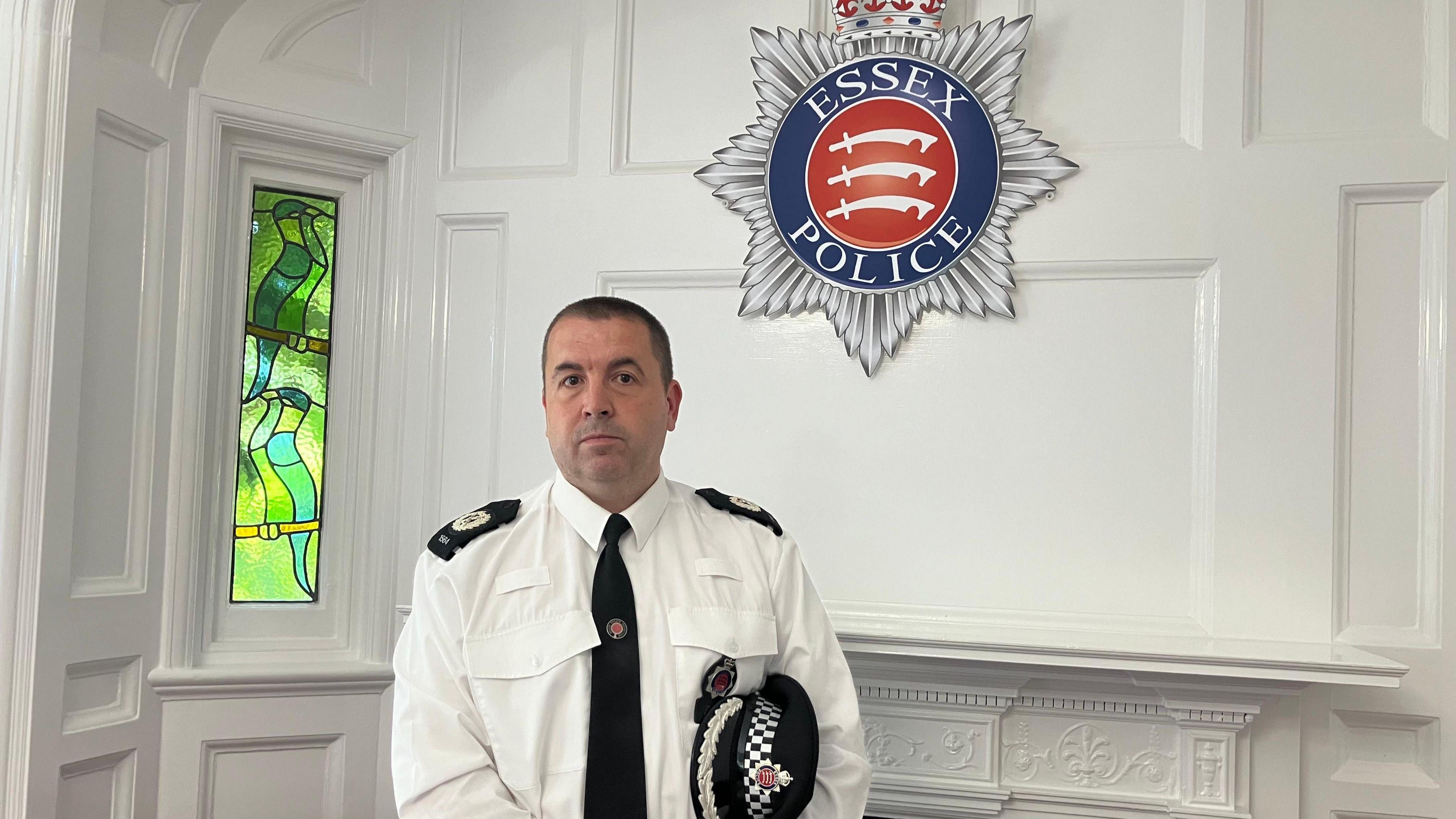 Stuart Hooper in a white shirt and black tie with a police hat under his arm and the Essex Police badge in the background