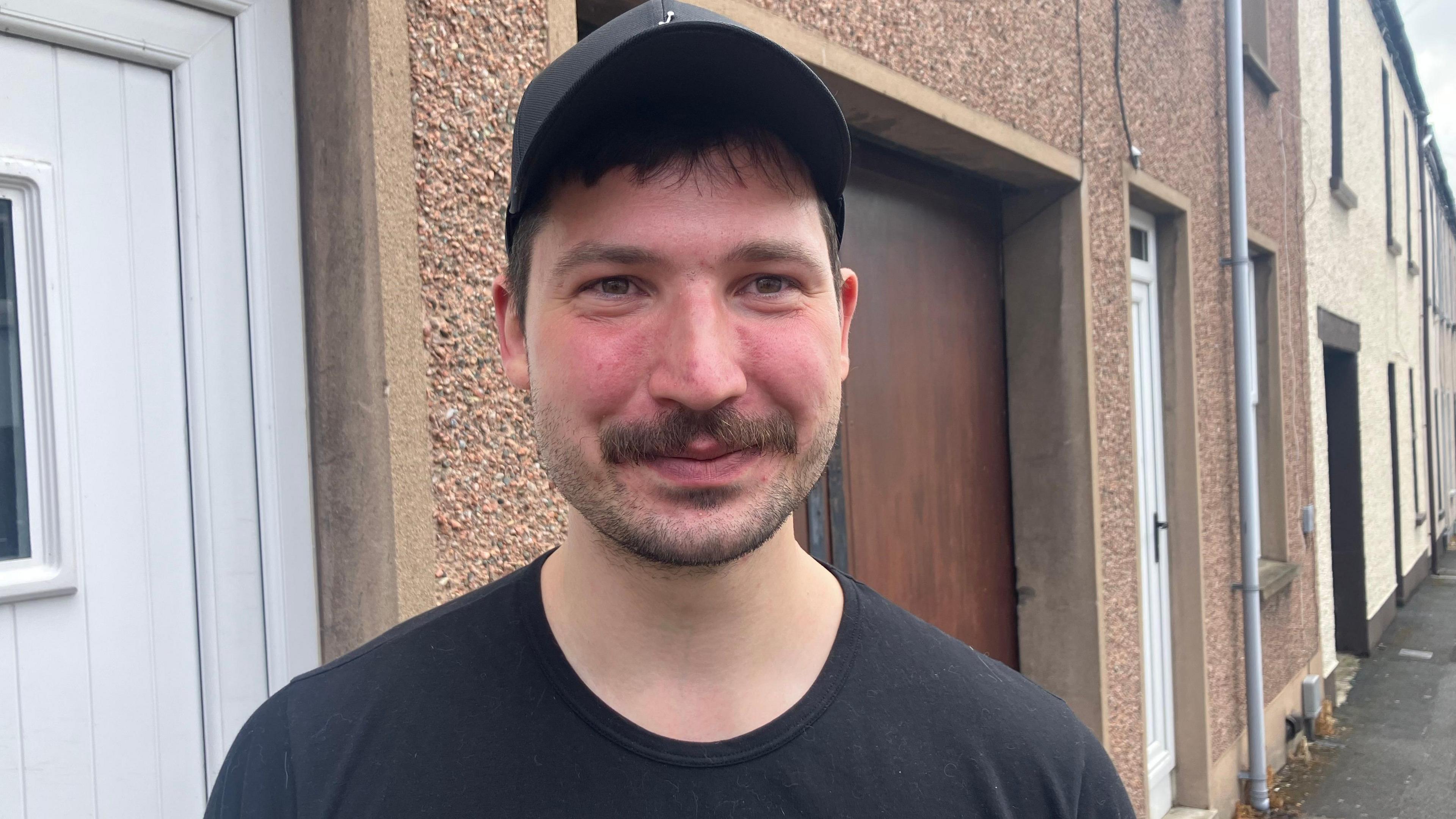 A man is smiling into the camera. He has brown hair and is wearing a black cap and a black top. He's standing on a street.