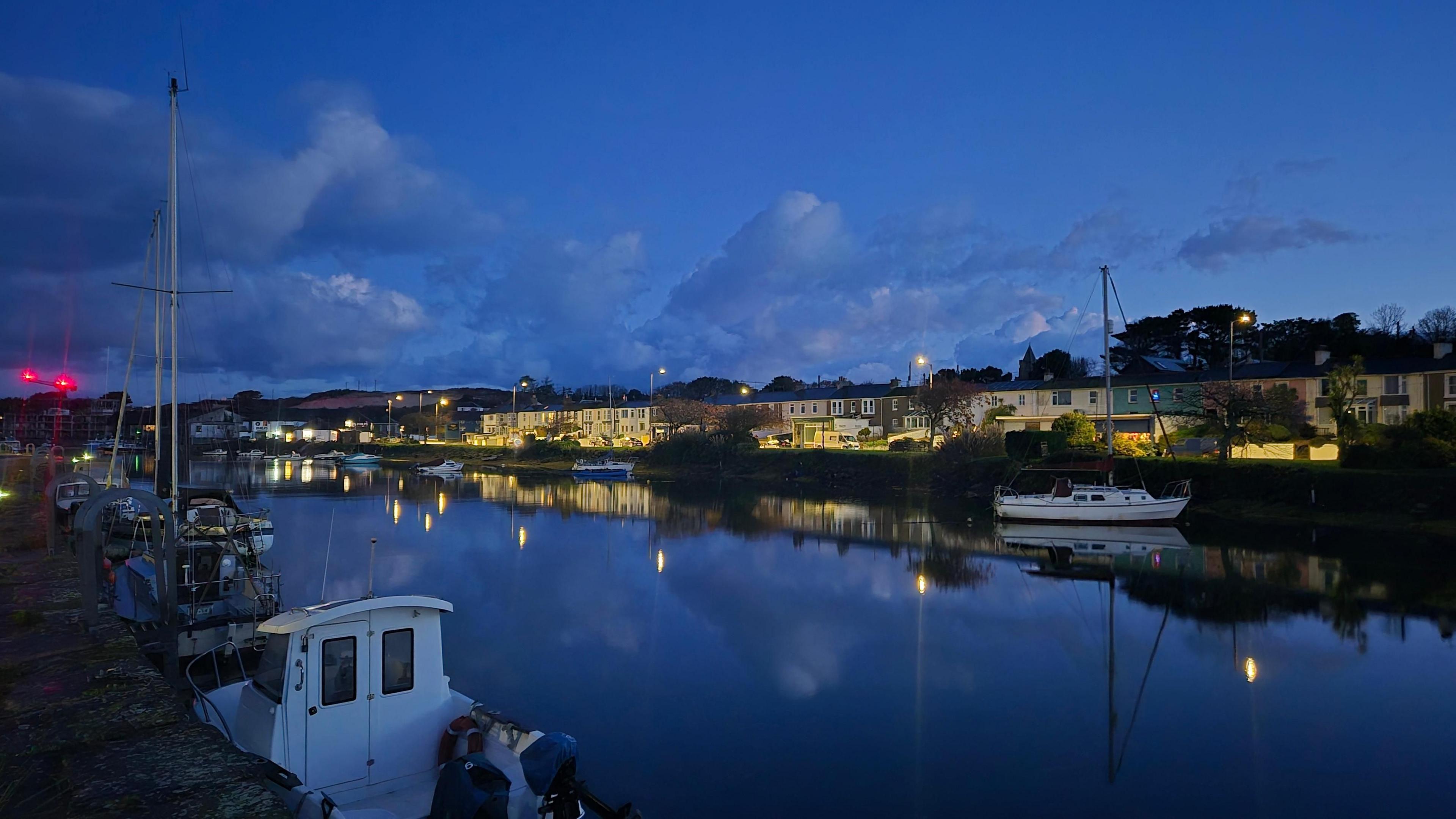 Houses line a body of water in Hayle, with street lights reflected in the water and boats seen in the foreground 