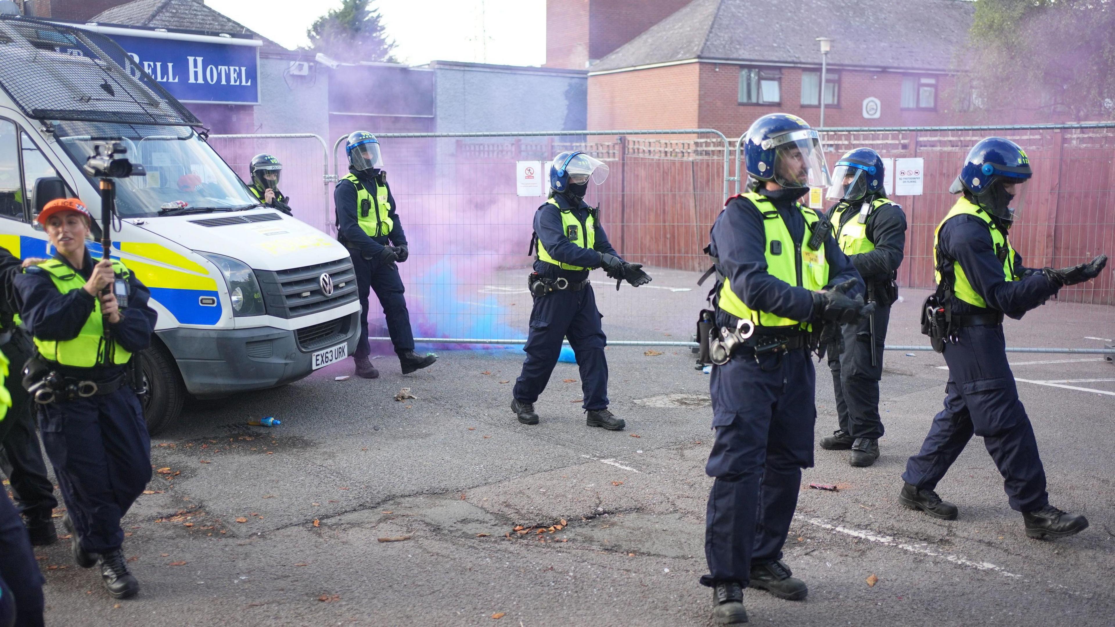 Police officers in riot gear standing in front of The Bell Hotel, which has its entrance protected by metal fencing. Blue and red flares have been thrown towards the officers and have smoke pouring towards the building.