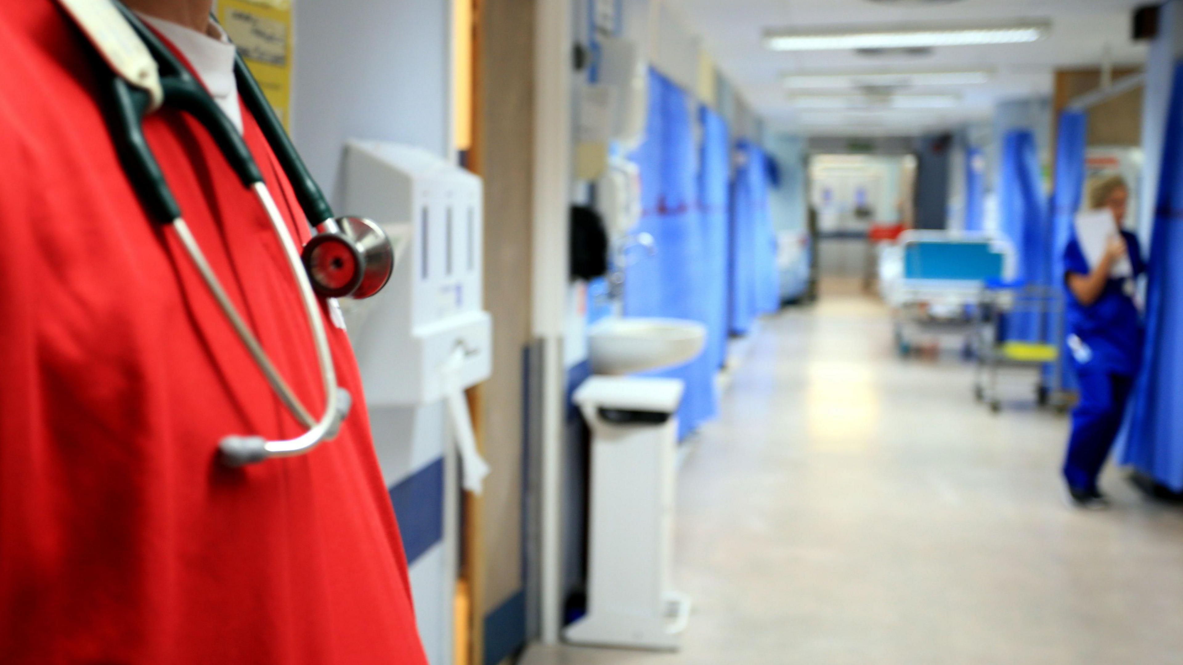 A hospital worker in a red tunic is in focus in the foreground of the picture standing to the left of frame. A stethoscope is hanging around his neck. A ward of bays with blue curtains around them stretches out behind him. A female nurse dressed in blue can be seen going into one of the bays. 