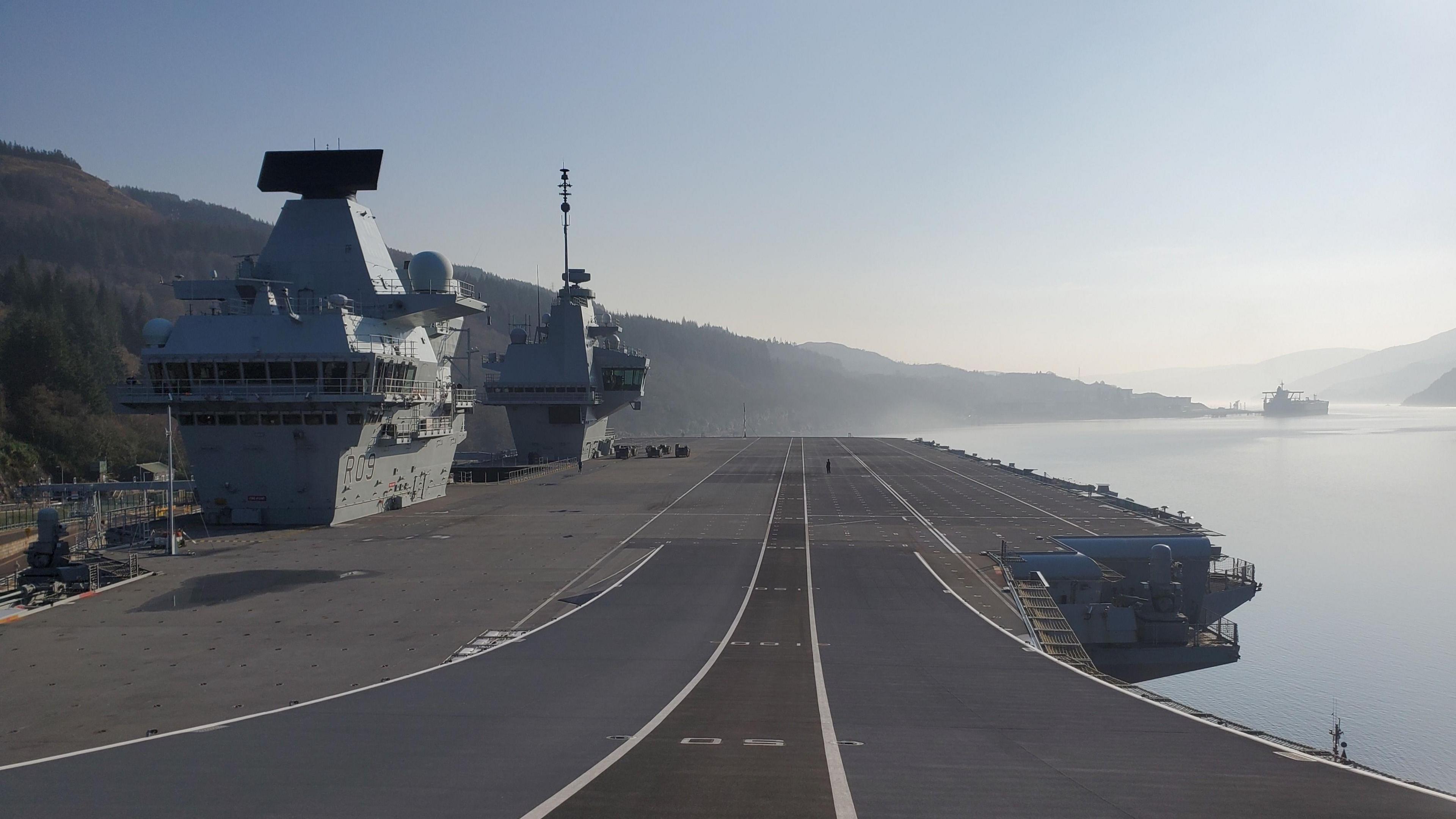 A general view of the flight deck on board the HMS Prince of Wales. The runway is grey with white lines on it. It is slightly curved in the foreground of the image leading towards the loch and mountains in the background. Two grey towers are on the left of the image.