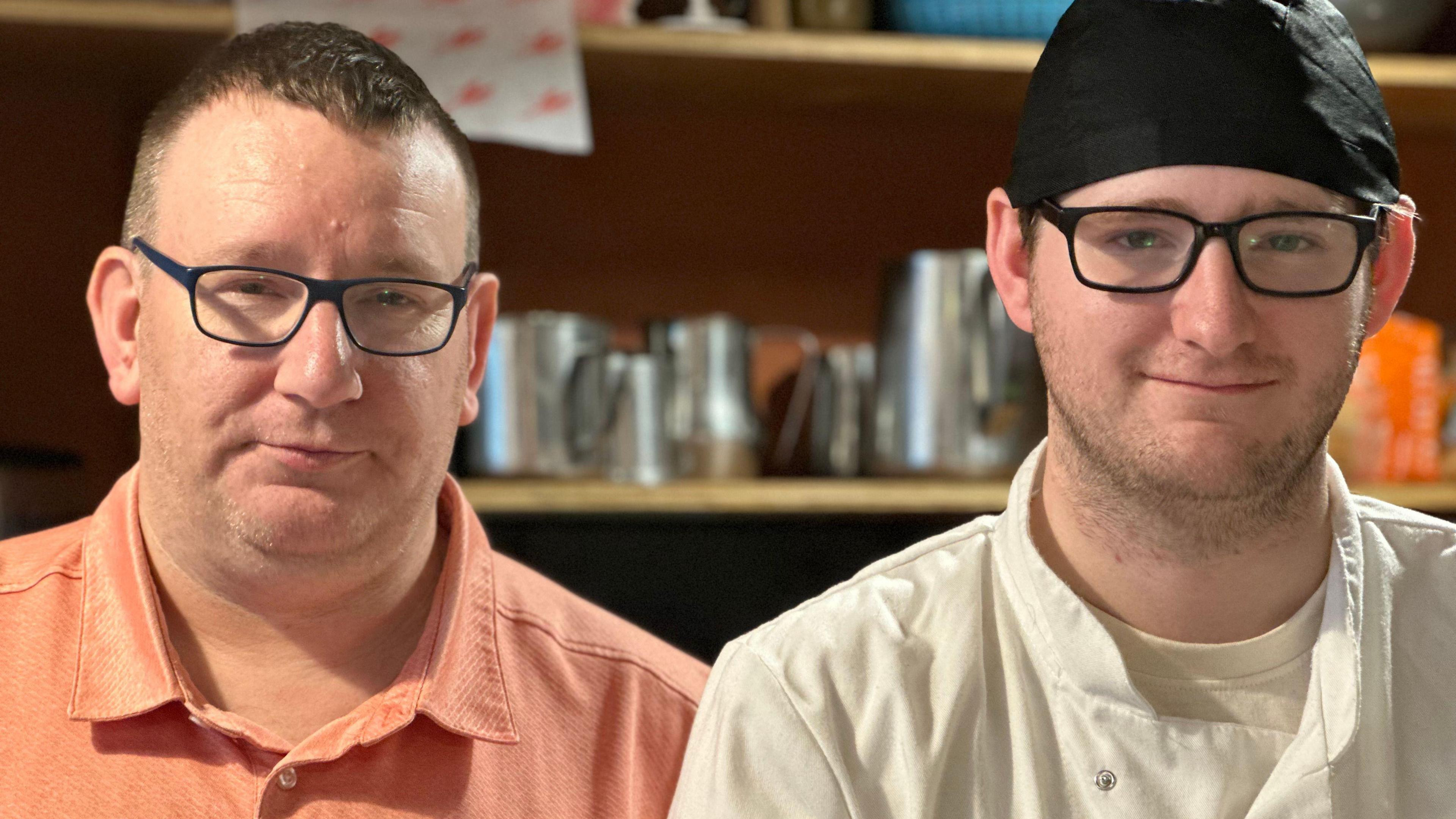 Ian and Jonathan Corlett standing next to each other, both wearing black rimmed glasses. Ian is wearing an orange T-shirt and Jonathan is wearing chefs whites and a black hat.