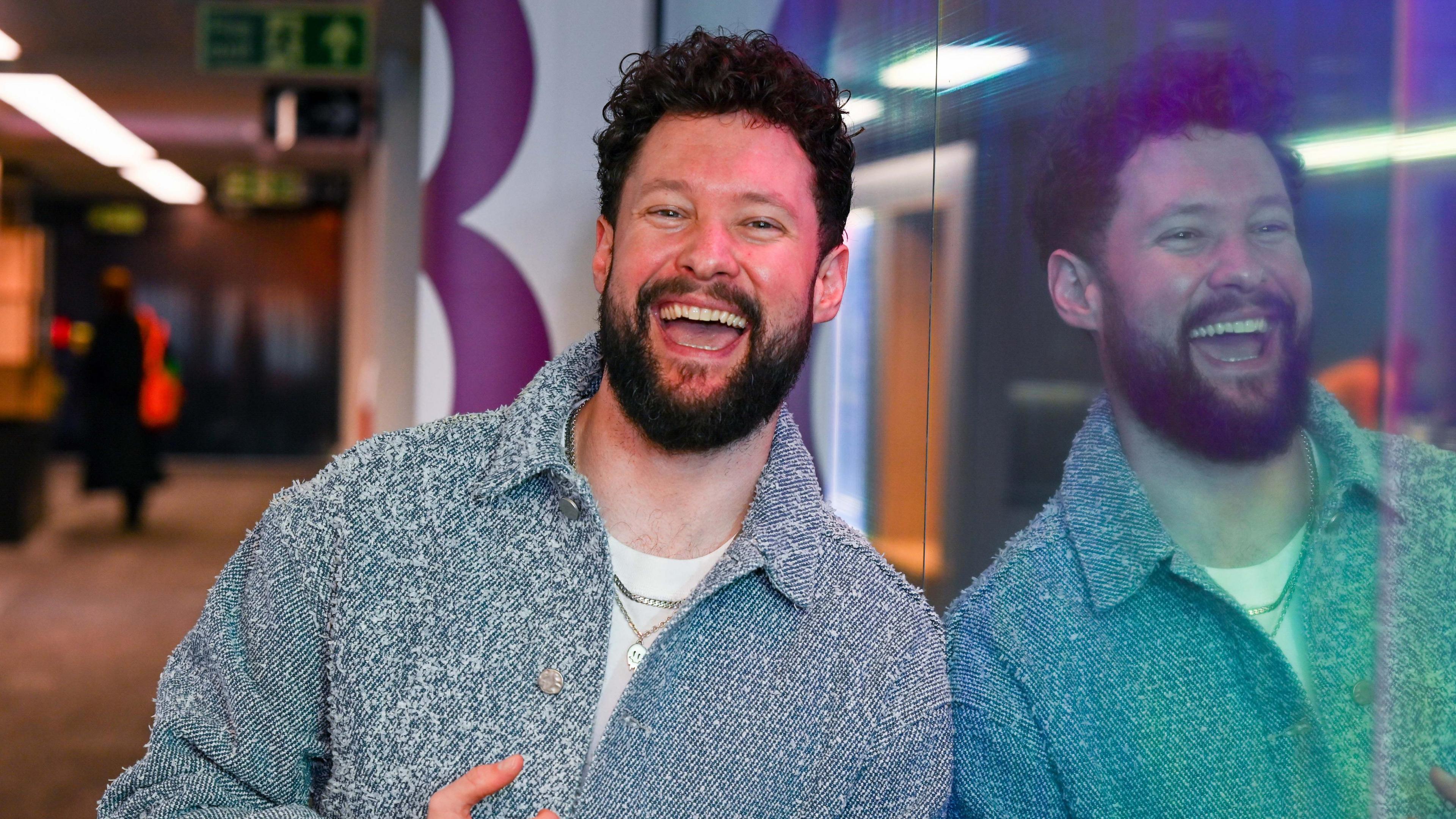 A man stands against a window in a radio studio with a broad smile on his face. He has curly brown hair, a brown beard and wears a chunky blue cardigan and white shirt. His reflection is clear in the window.