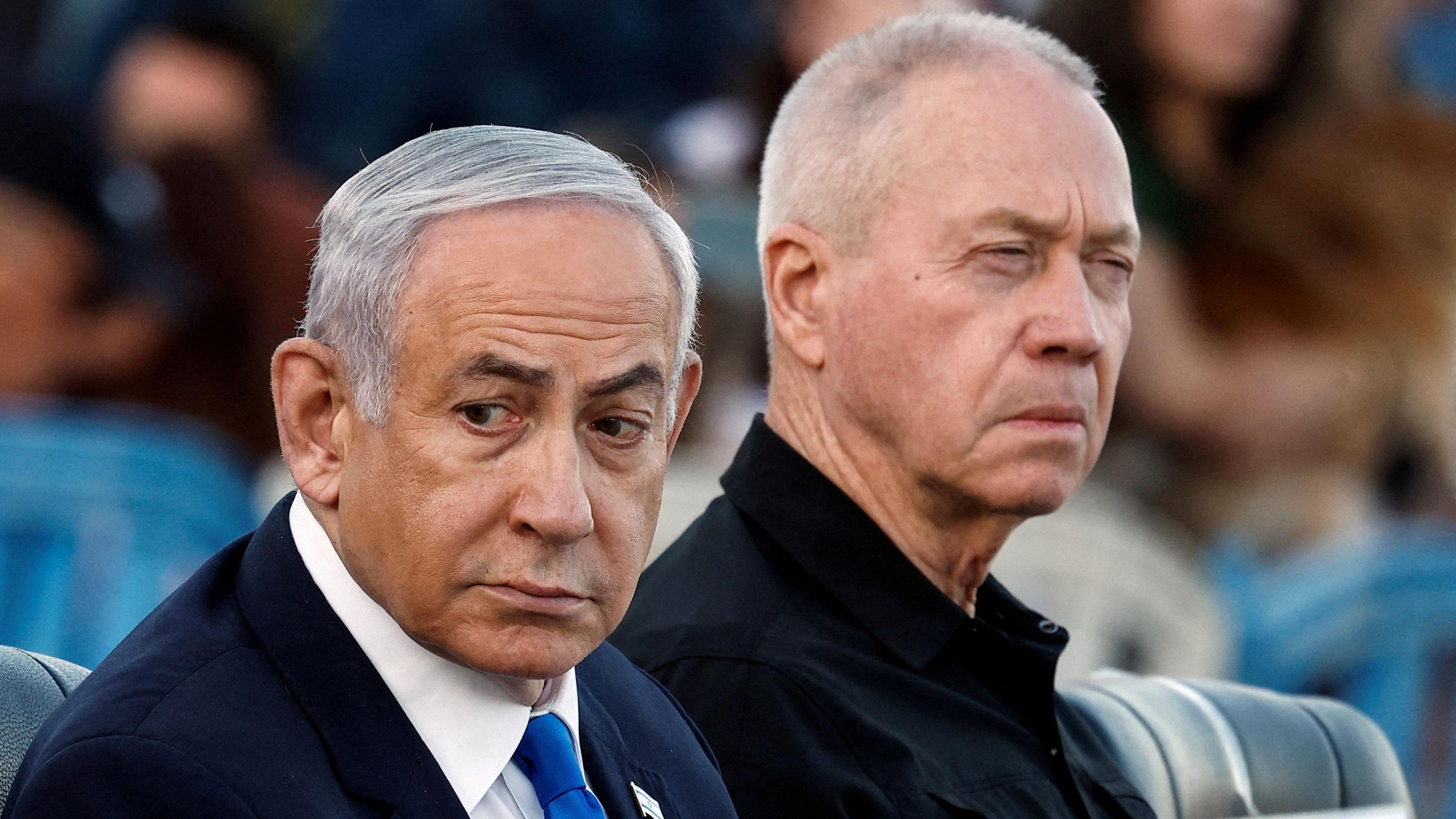 Benjamin Netanyahu, with combed over grey hair, wears a black suit, white shirt and blue tie and looks over his right shoulder as he sits next to Yaov Gallant who is wearing a black shirt and squinting