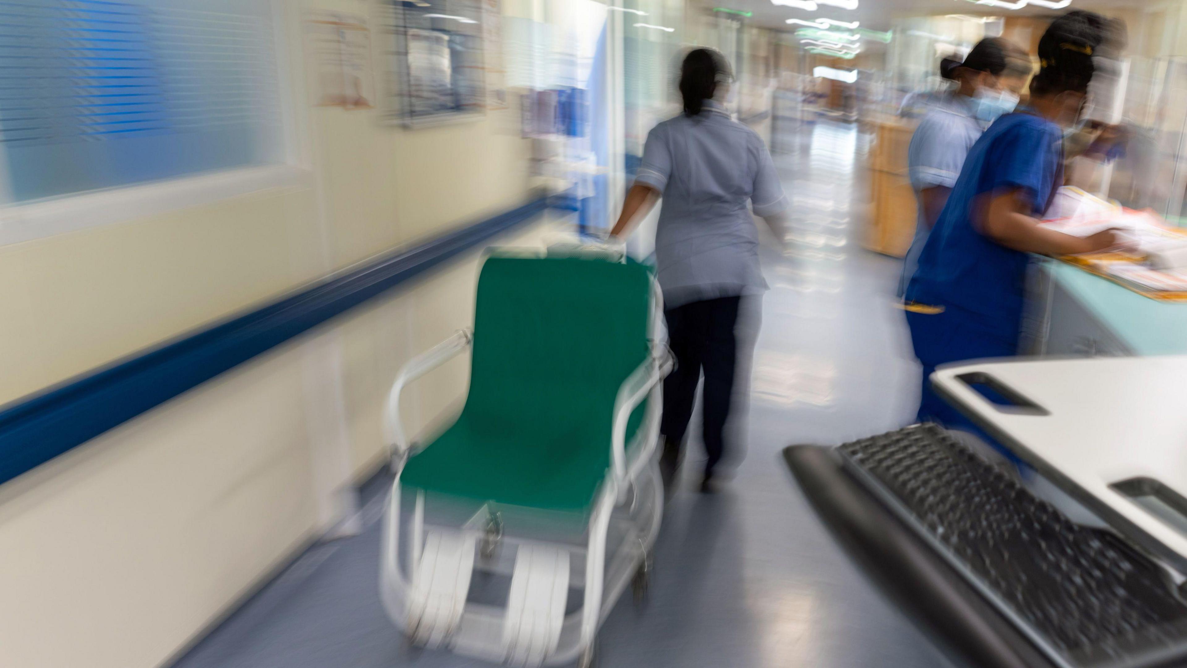 A large green and white wheelchair being moved at speed down a hospital corridor by a member of staff while two others stand nearby looking at notes. There is a black computer keyboard in the foreground. 