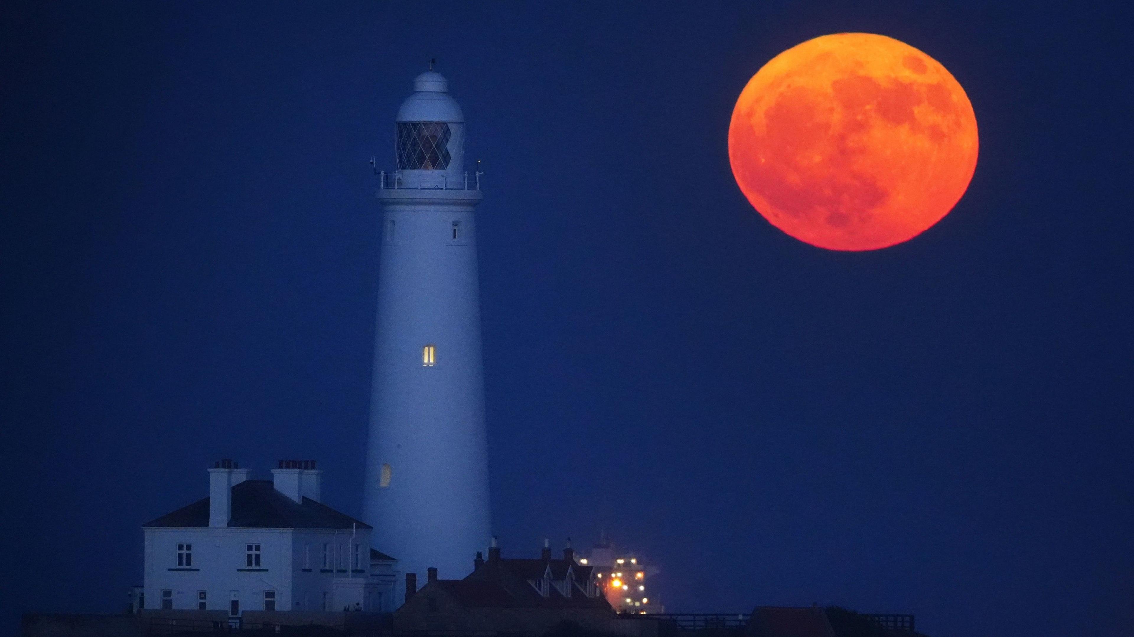 A red-orange full Moon hangs low in the night sky next to a lighthouse