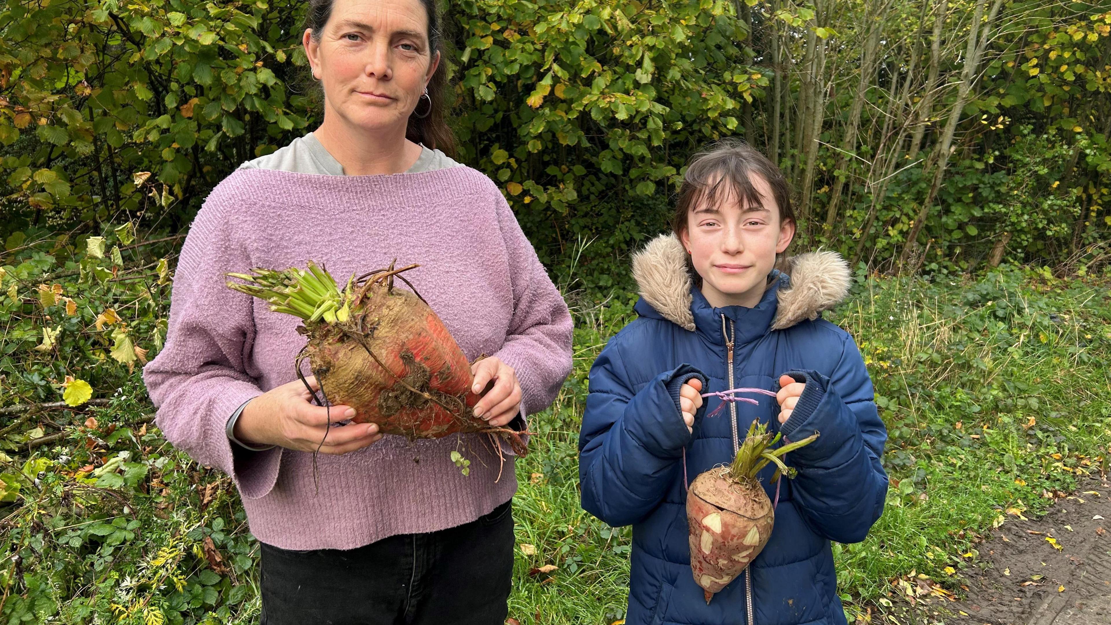 Dawn Qince, with brown hair and wearing a purple jumper, is holding a Mangelwurzel to carve and and her daughter Cherry, wearing a blue coat, is holding a carved lantern. They are both stood outside with bushes in the background.