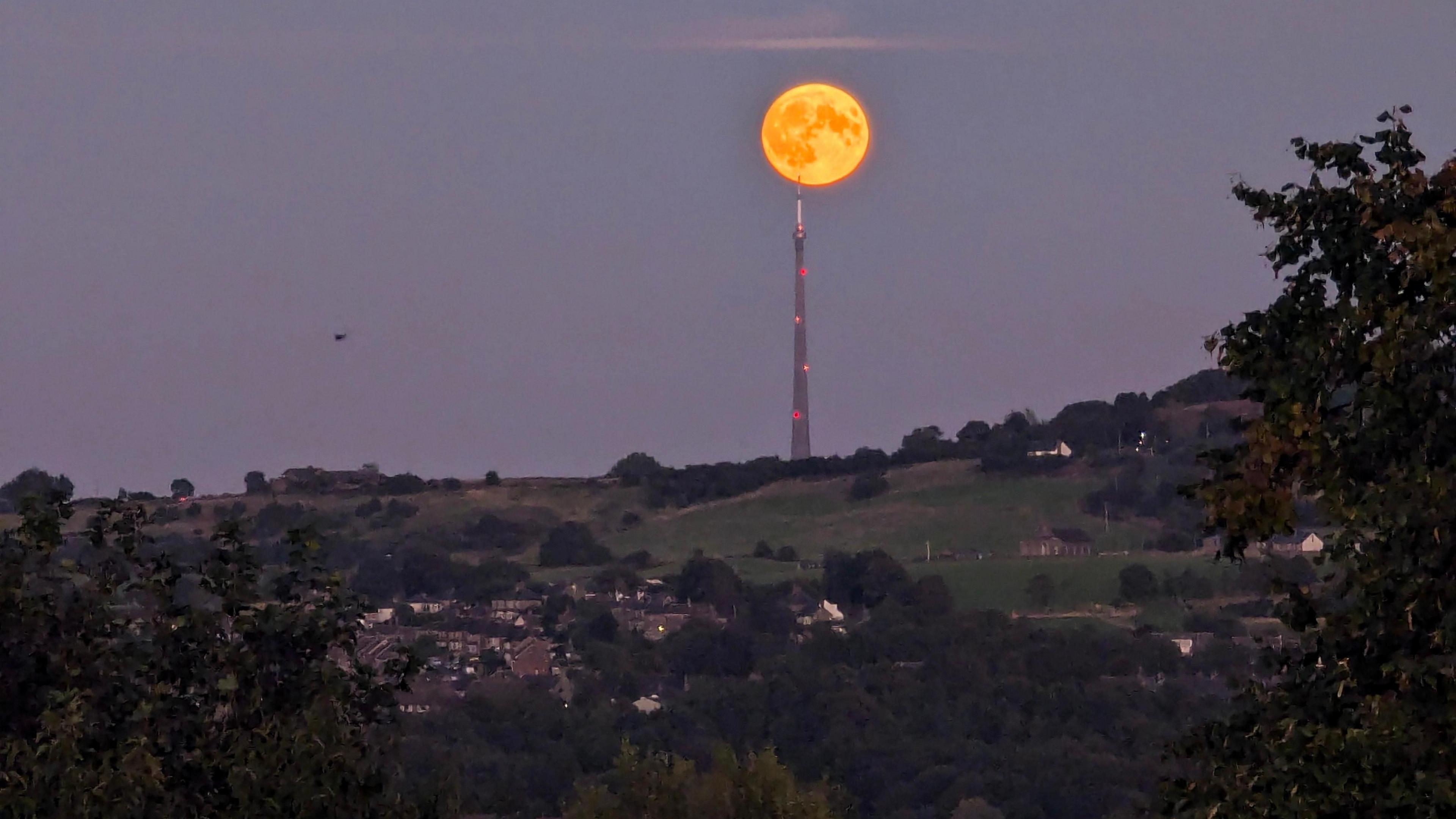 An orange glowing moon above the Emley Moor transmitting station in Huddersfield. Several houses are dotted along the rolling, green hills. 