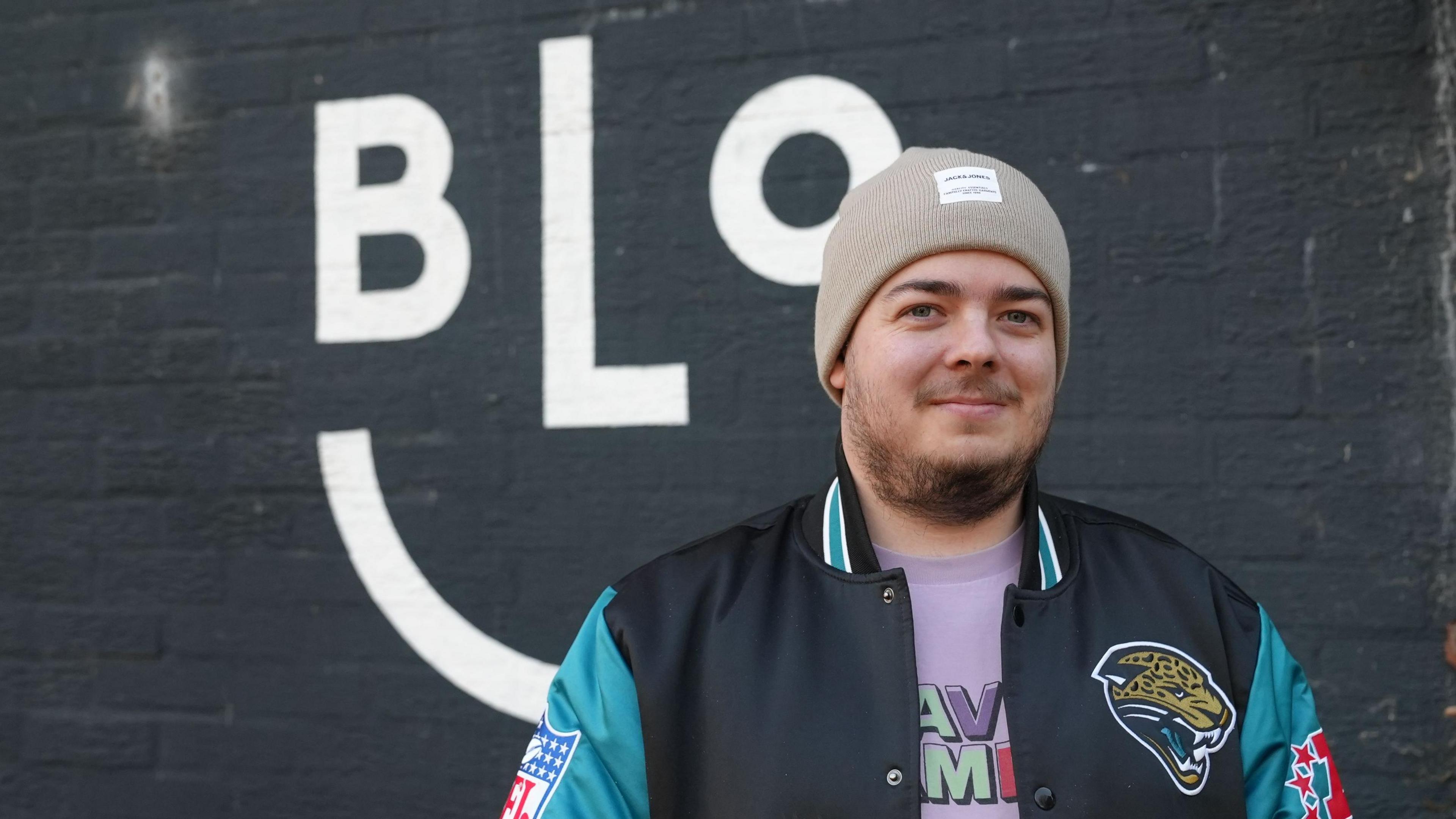 Photograph of Elliot, the owner of Bloc coffee shop in Victoria Park, along Cowbridge Road East. Elliot wars a navy and blue bomber jacket, a pink graphic t shirt and a beige beanie hat. He has blue eyes, a dark brown beard and smiles at the camera. 