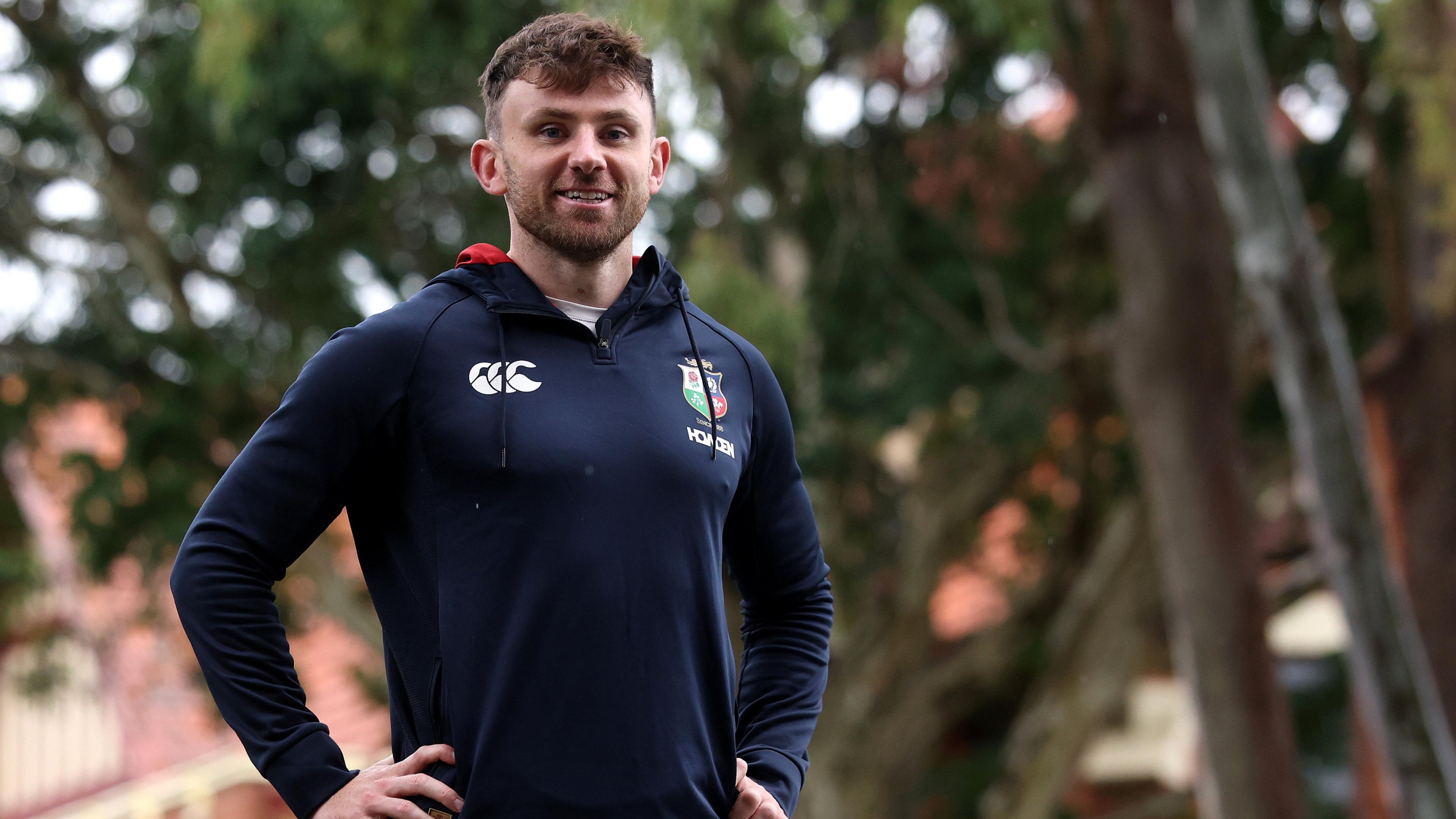 Hugo Keenan stands with hands on hips and smiles - there is a tree out of focus in the background