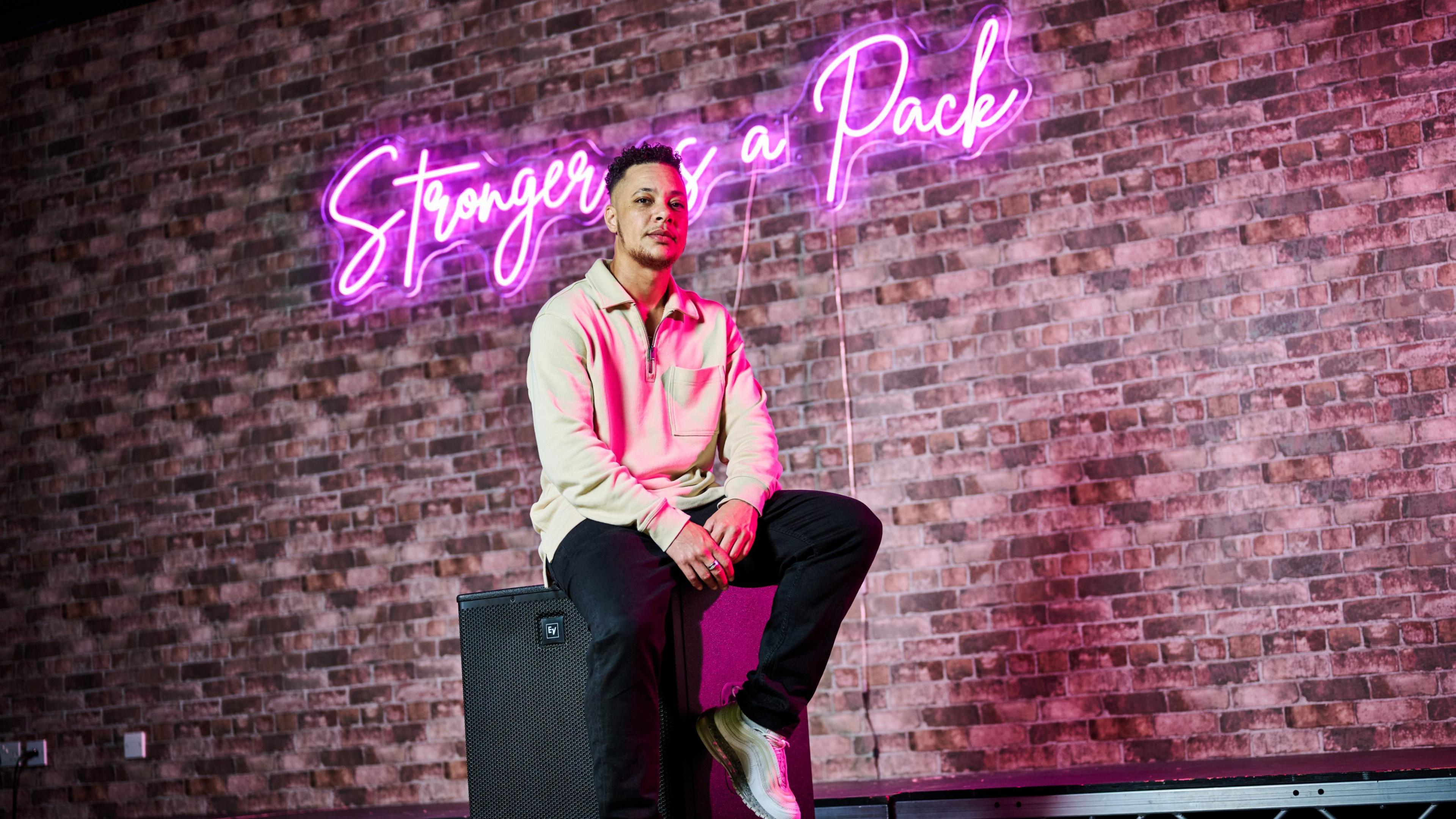 Nate Ethan-Watson is dressed in a beige long-sleeved top and black trousers with trainers. He is sitting on a black speaker in front of a red-brick background. On the wall is a neon pink light that reads "Stronger as a pack." the pink light is reflecting on his face and clothes.