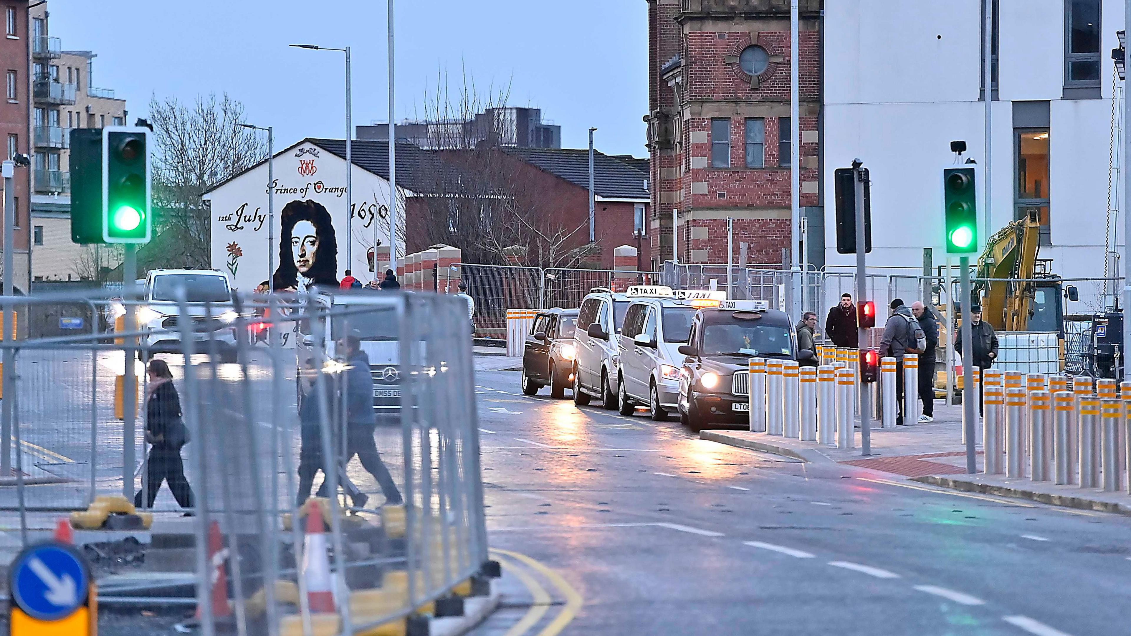 The road reopened - taxis are pictured lined up parked at the side of the road. There are people pictured on the pavement behind some traffic lights. On the left of the photo, there are construction fences.