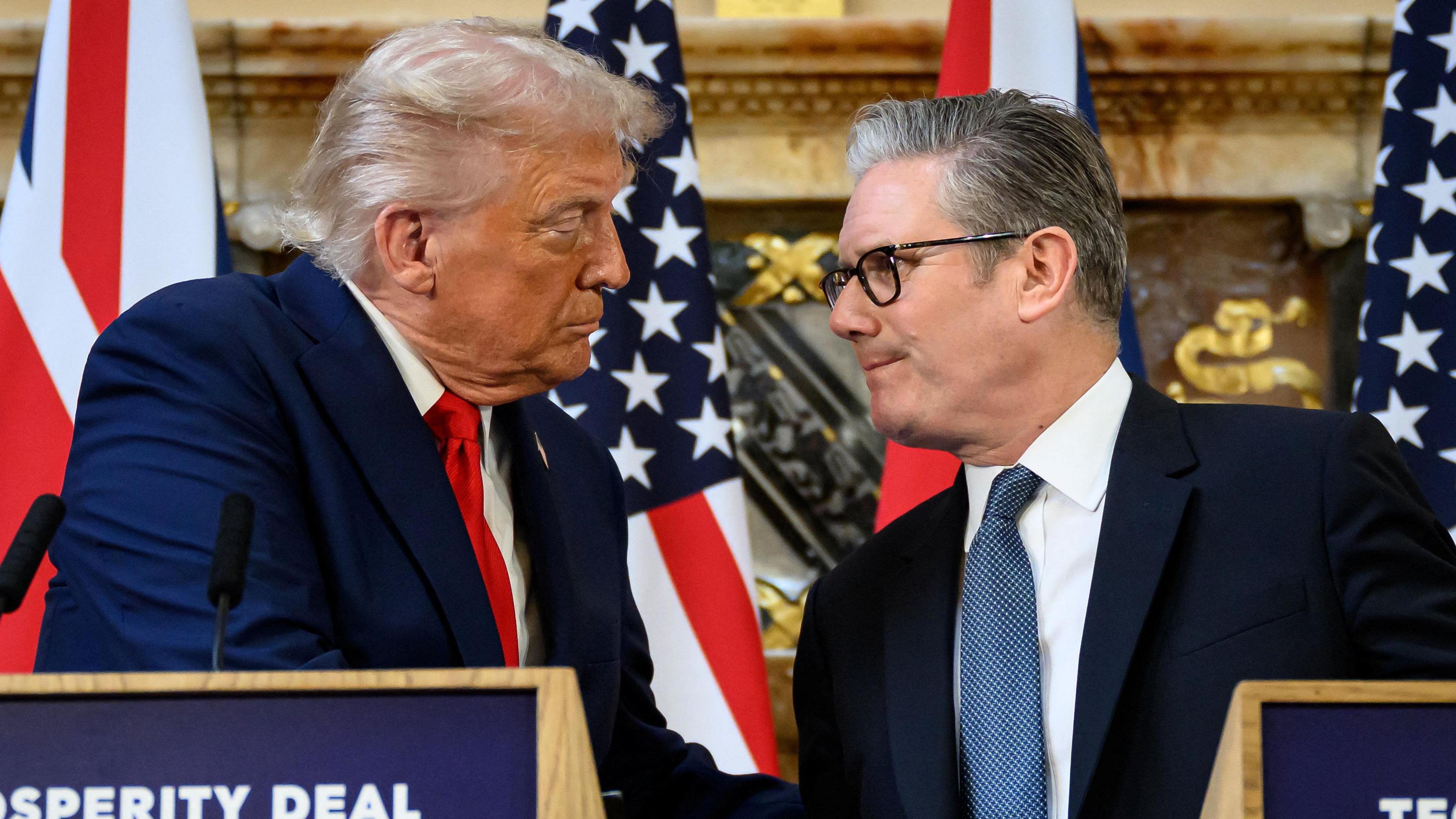 US President Donald Trump and Prime Minister Sir Keir Starmer shake hands with US and UK flags behind them. They stand in front of wooden podiums.