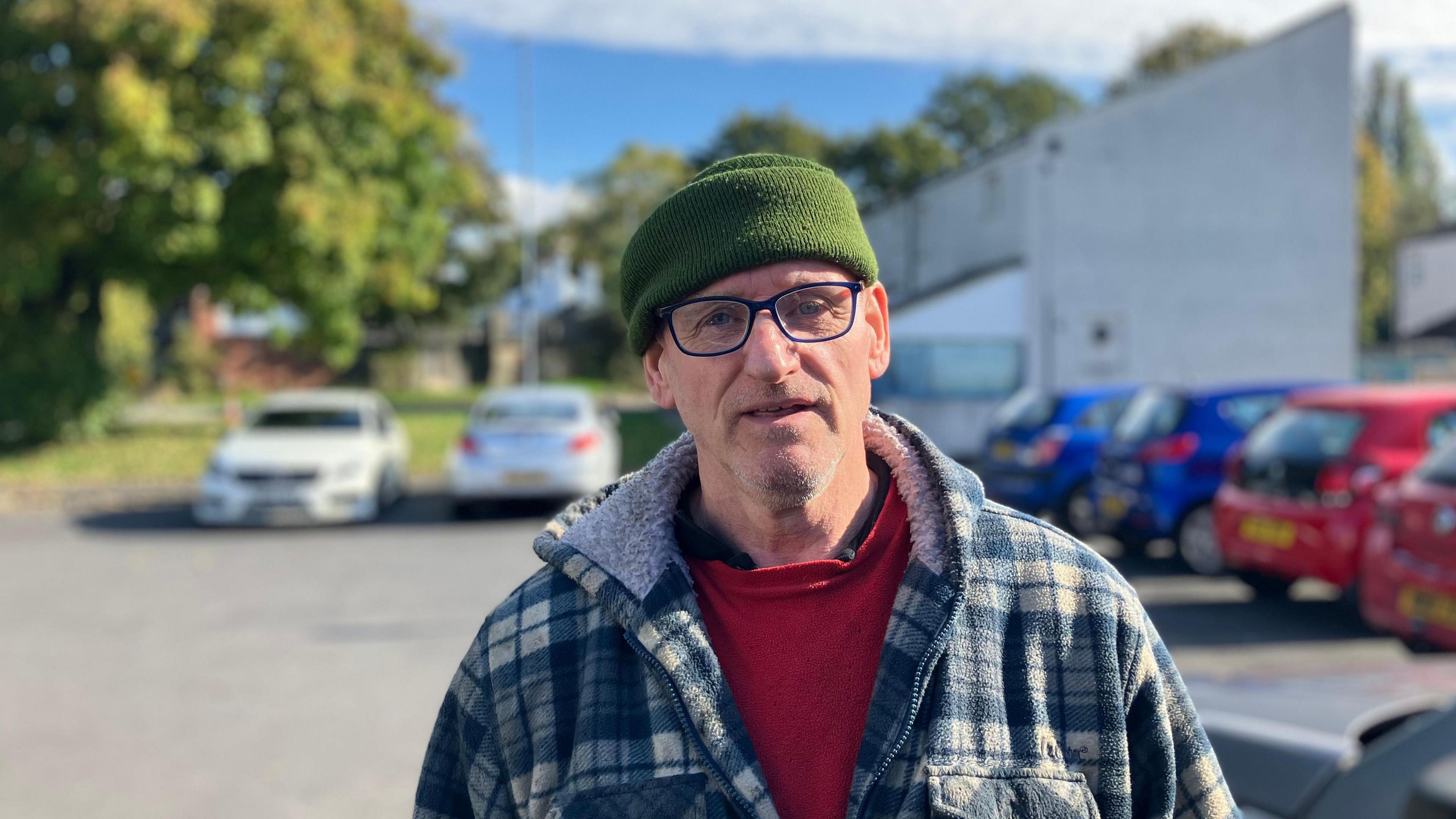 A man with a green hat and blue glasses wearing a fleecy blue coat and standing in a car park