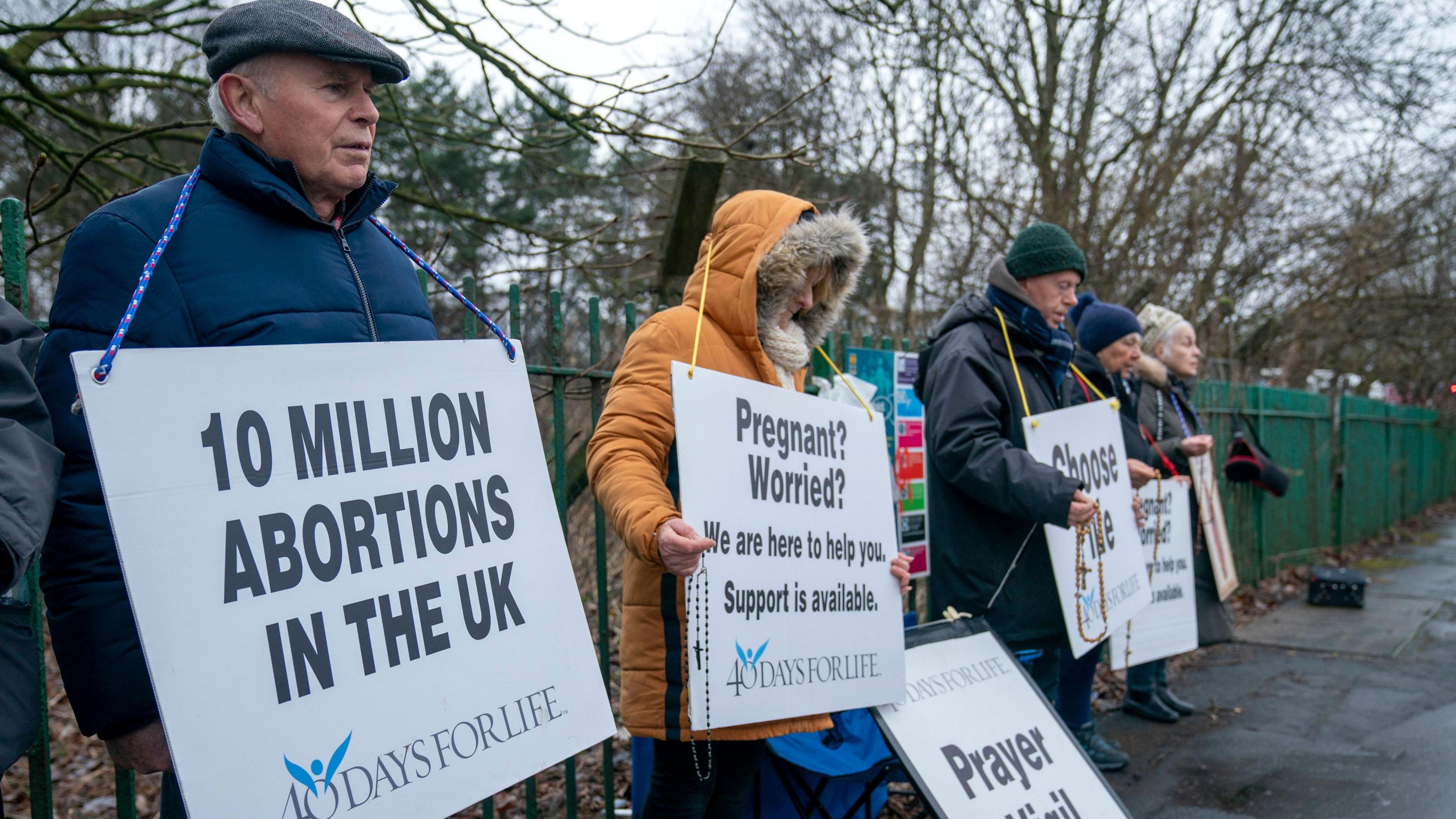 Anti-abortion protesters holding placards near a hospital in Glasgow. Five demonstrators are pictured. They are all wearing warm winter clothing and some are holding rosary beads.