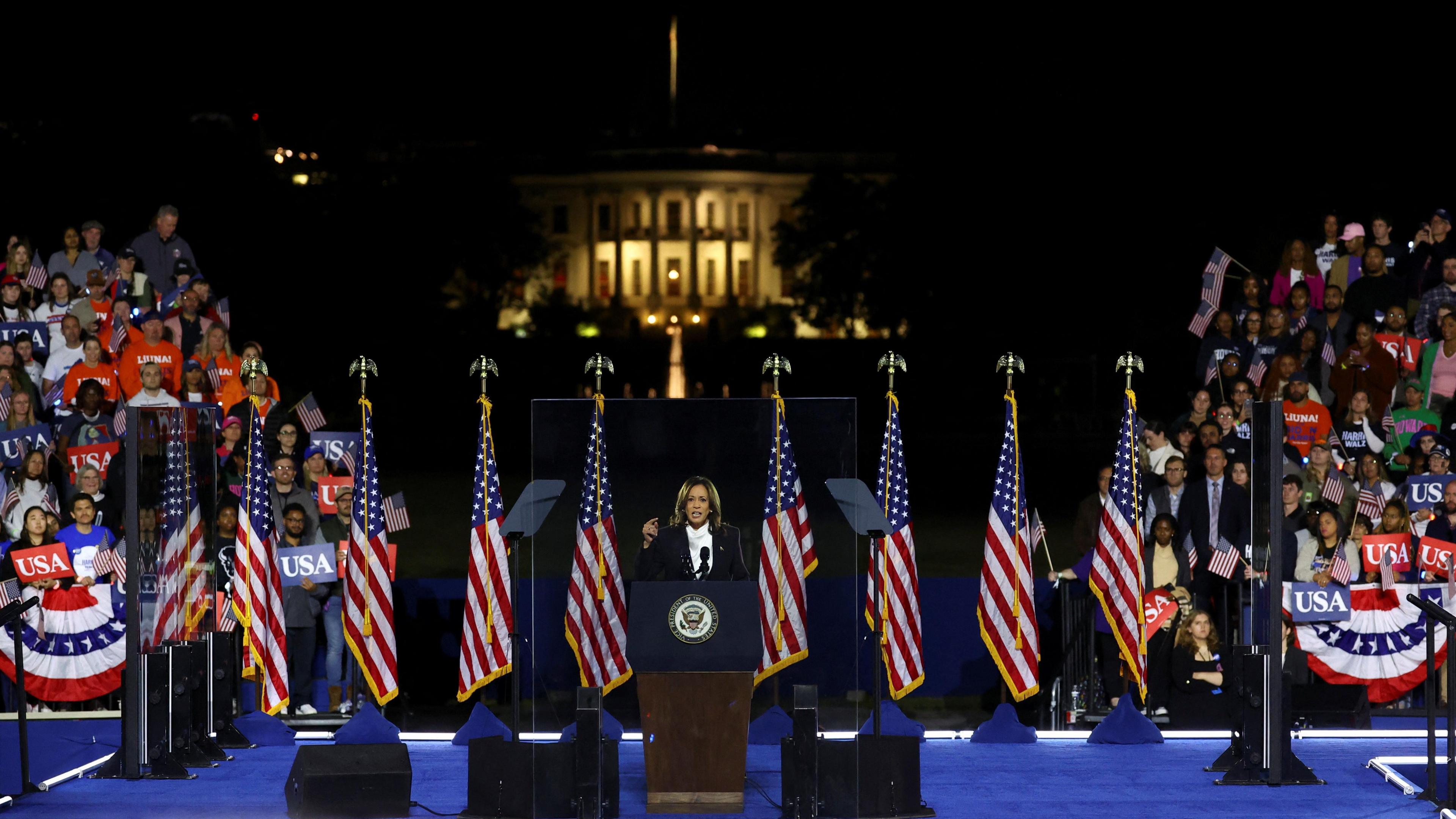 Vice President Kamala Harris stands an a podium at night on a blue stage with American flags behind her and the White House lit up behind her. There are some crowds to the right and left behind her.