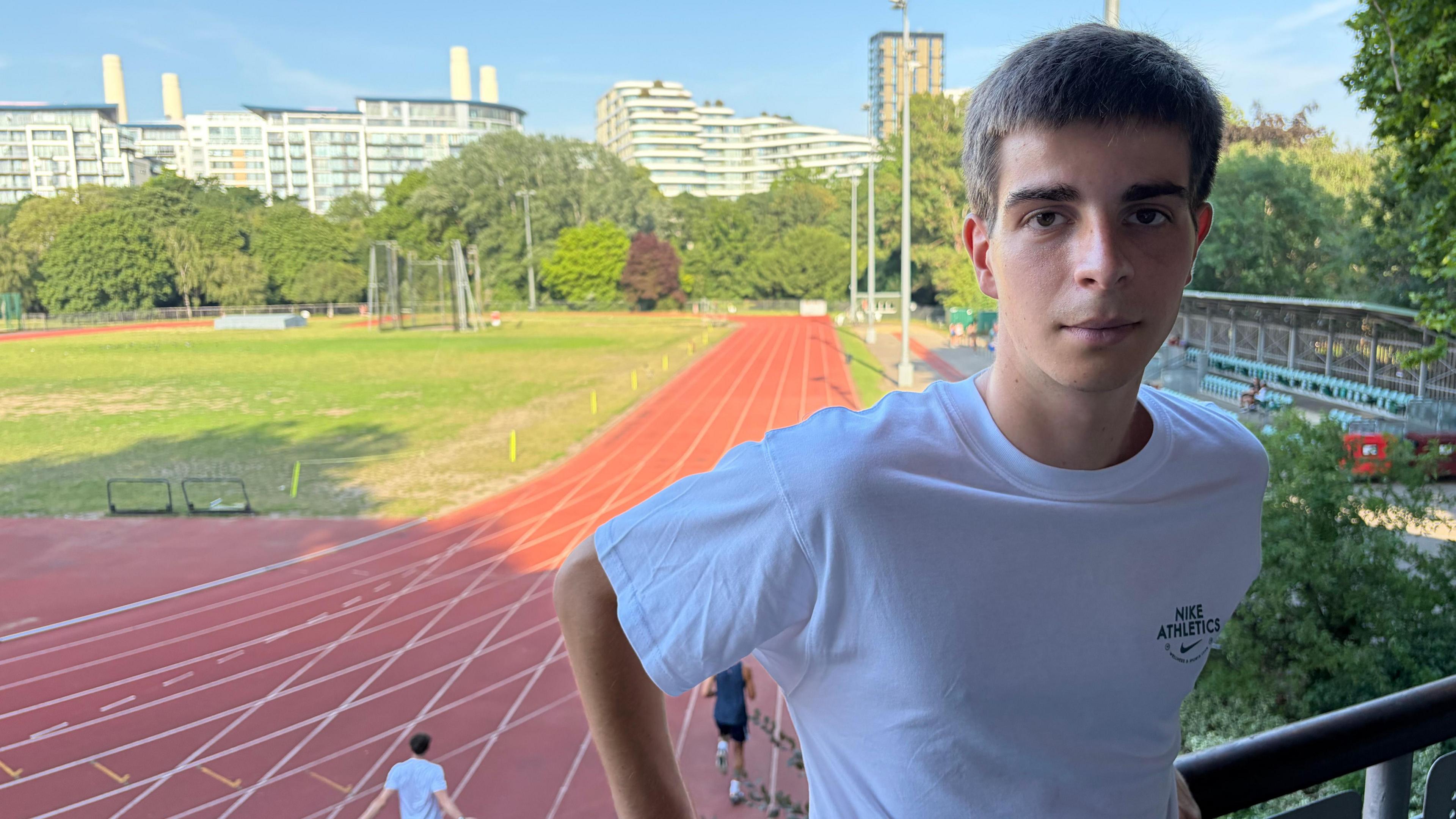 A man stands on a balcony overlooking an athletics track, wearing a white T-shirt.