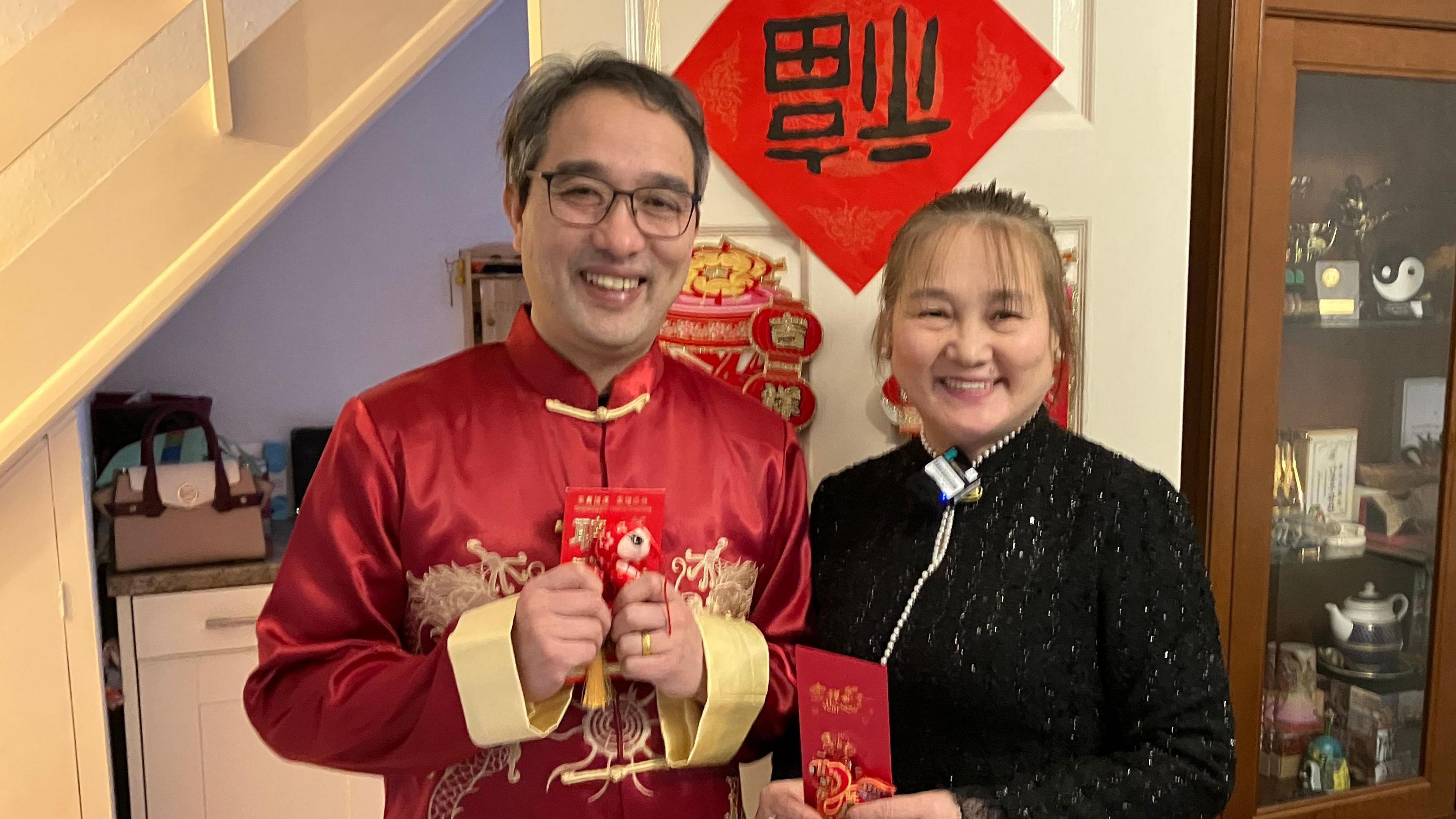 Tie Chen, wearing traditional red and gold Chinese dress next to Amy Zhen who is wearing a black sparkly top and both are standing in front of a glass-door display cabinet. 