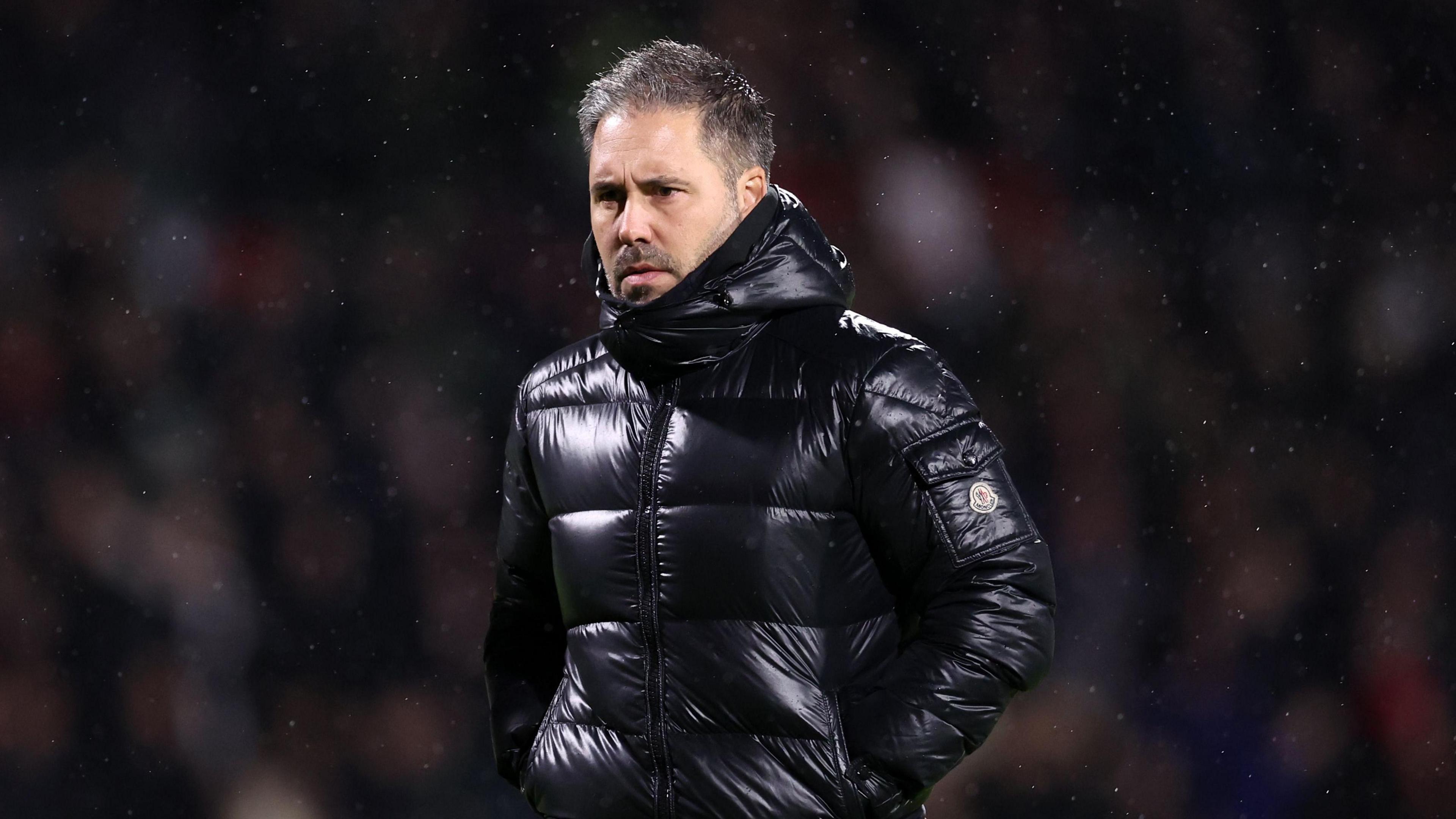 Marti Cifuentes watching his Leicester City side face Wrexham, standing in the rain wearing a shiny, black puffer jacket.