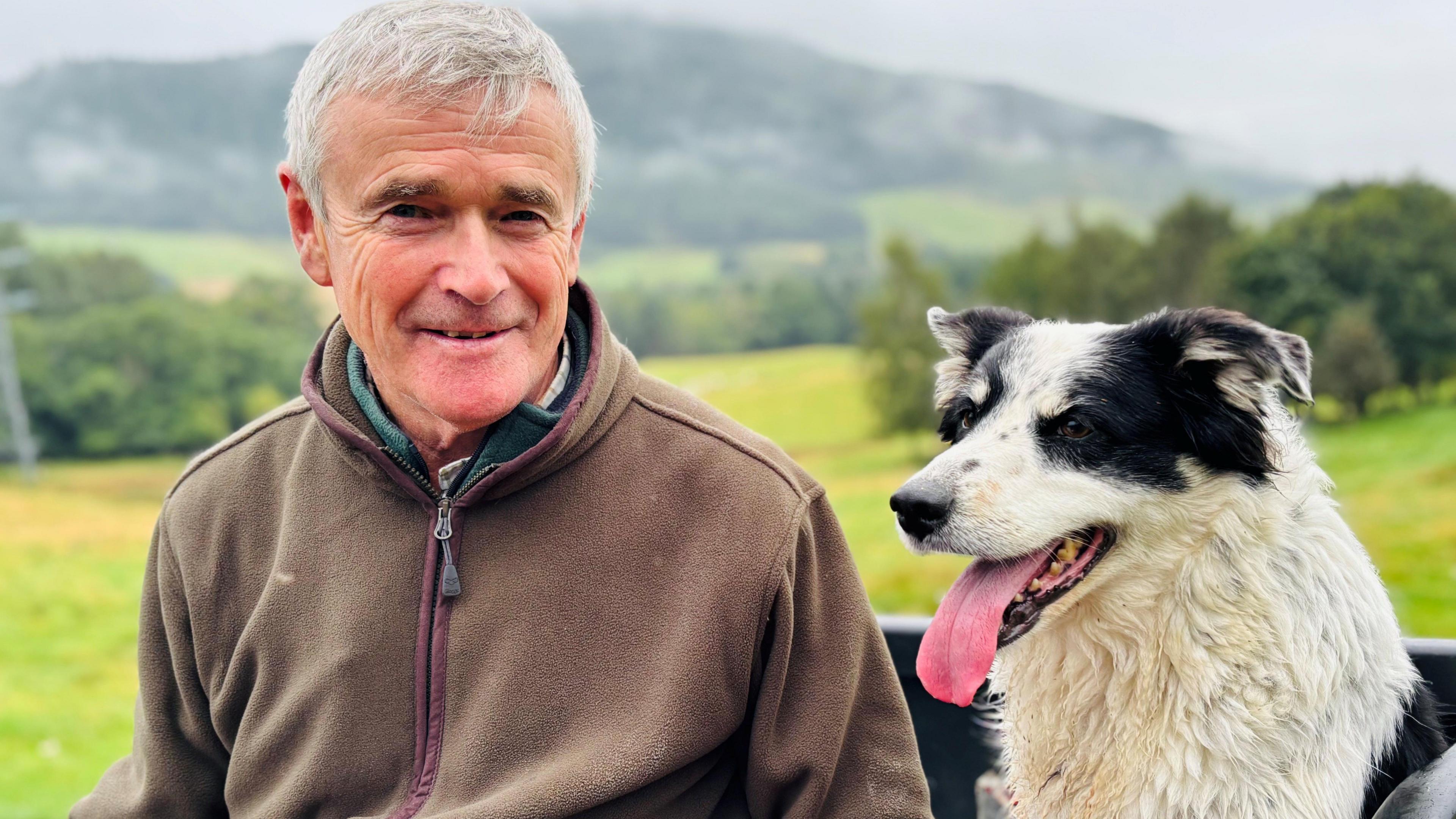 Farmer Ian Duncan Millar, smiling at camera, sitting next to a black and white sheepdog with its tongue hanging out, with fields, trees and hills in the background.