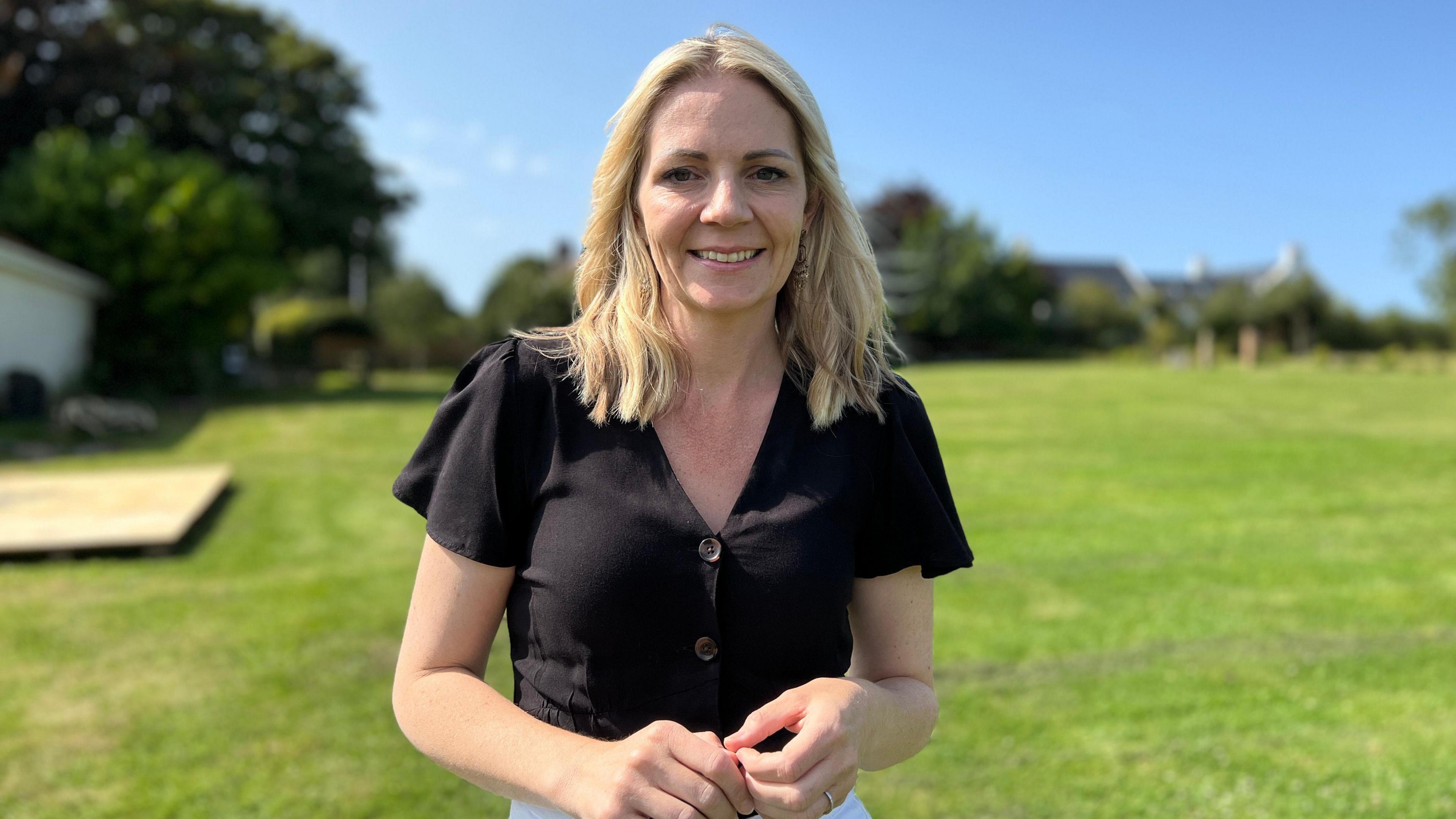 Chloe smiles at the camera with a field in the background. She is wearing a black shirt and she's holding her hands. Her blonde hair is being blown slightly in the wind.