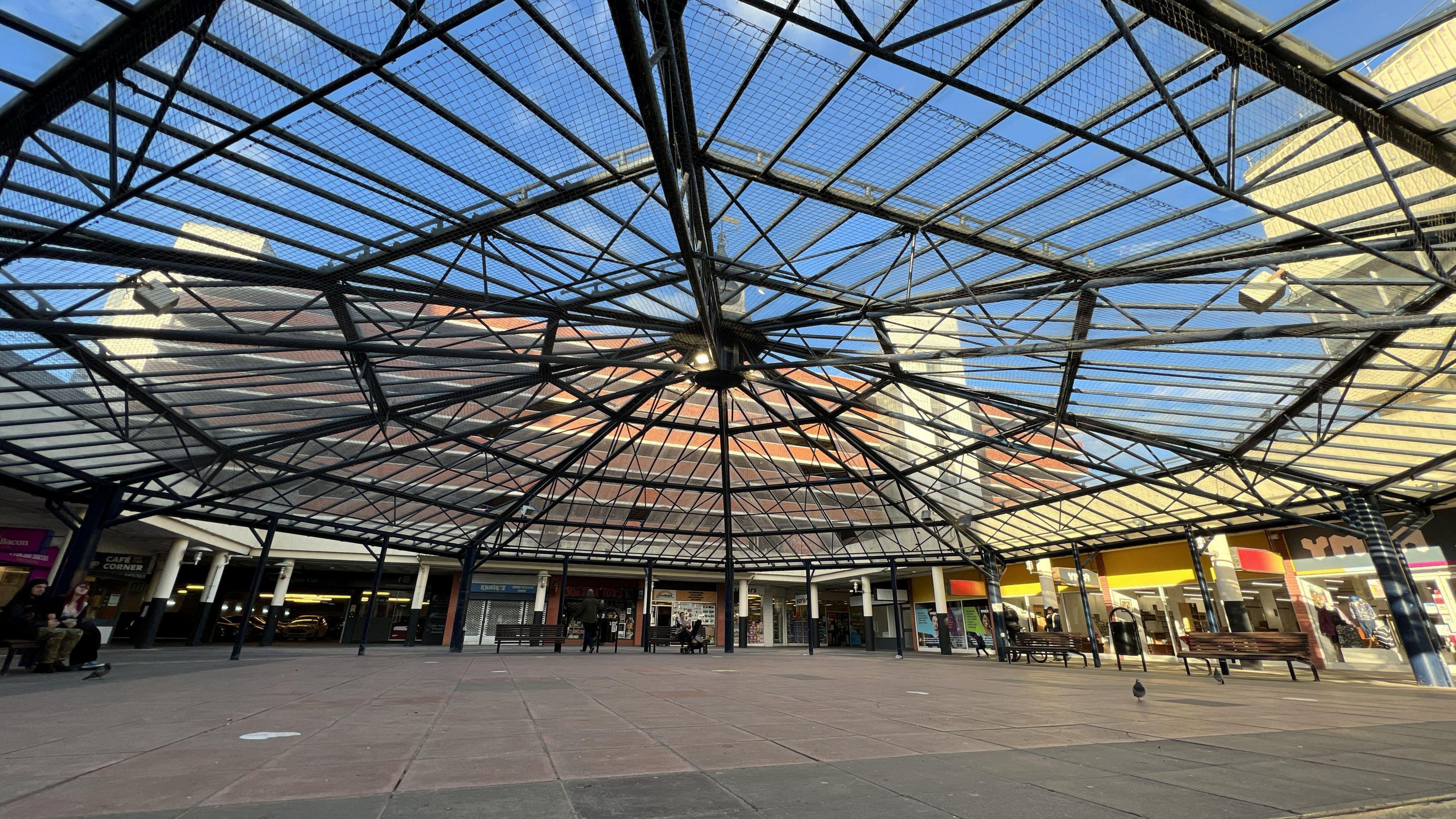 A picture of the central precinct area of Anglia Square. It has an octagonal metal framework covering it, with various shops around the edges.