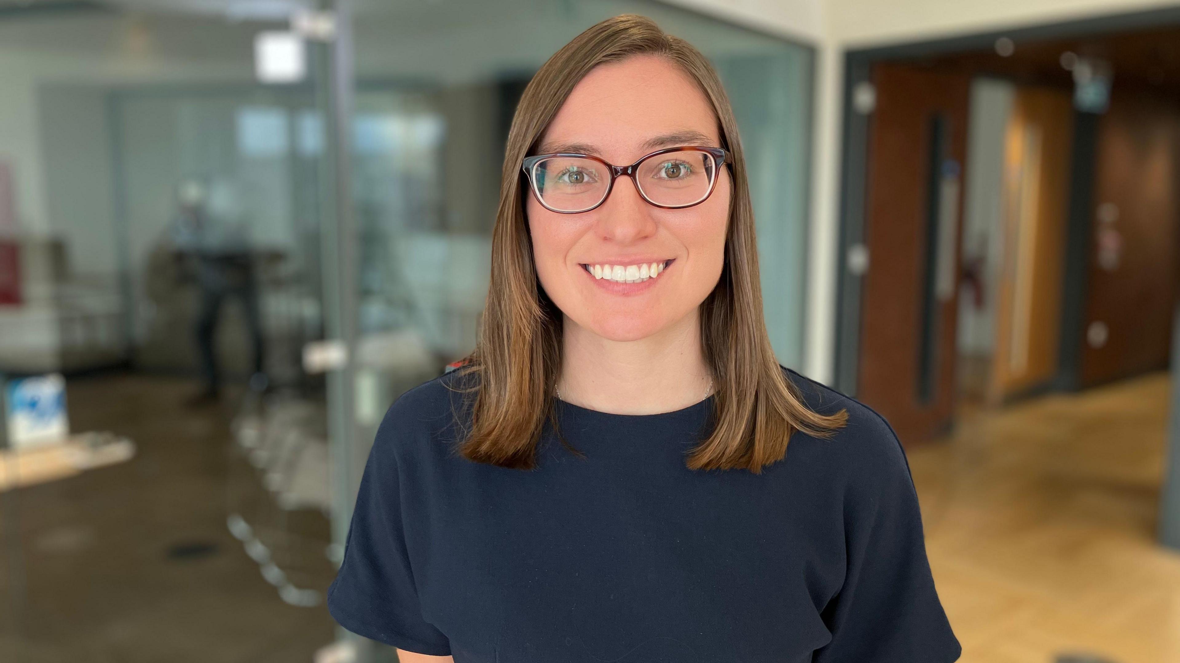 A head and shoulders shot of Sarah Hayward who is looking at the camera and smiling. She is wearing glasses and a blue top. The background is blurred but she appears to be standing in a corridor with some wood panelling behind her and a glass fronted office.