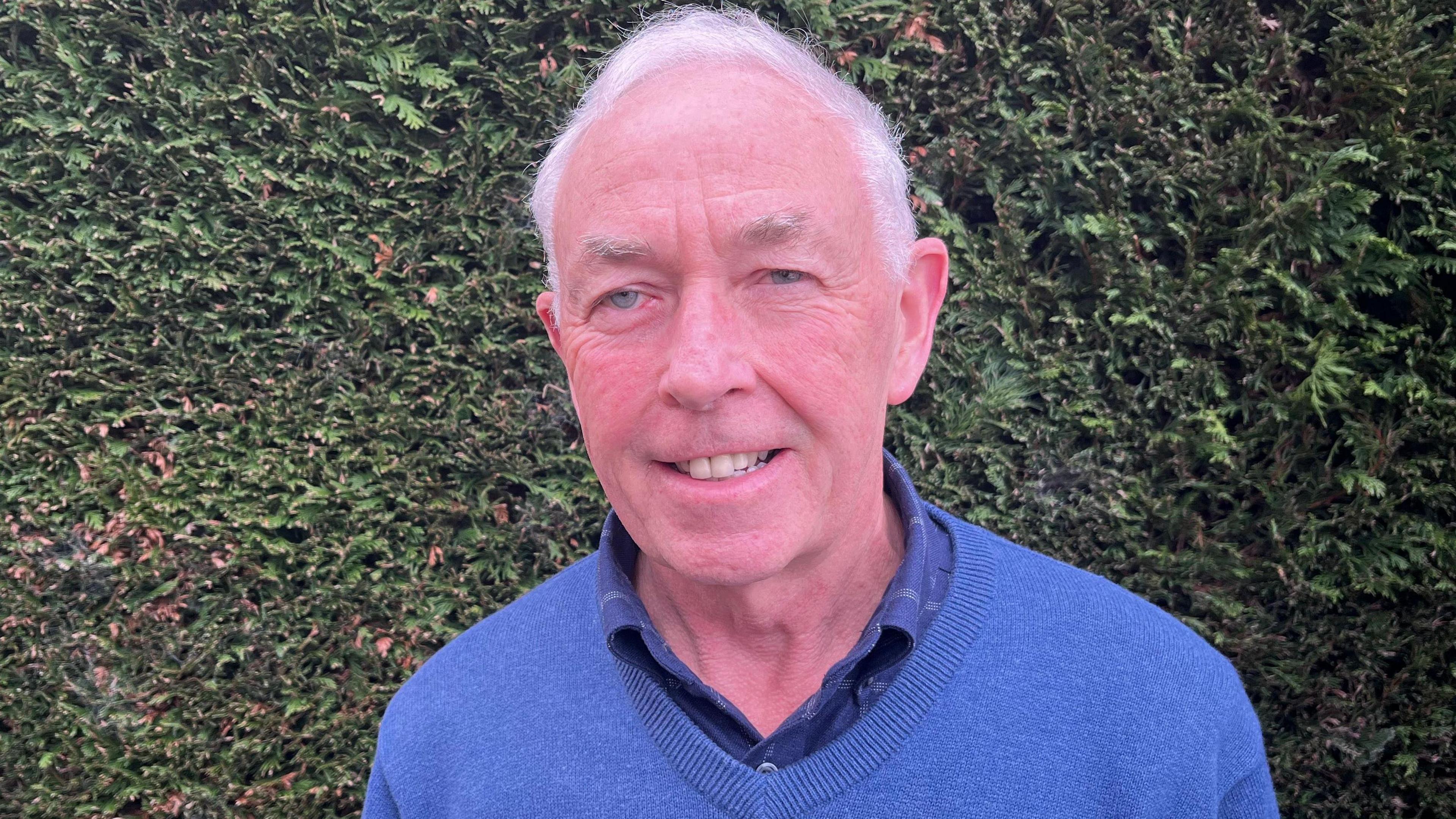 Pete Russ smiles at the camera, standing in front of a hedge. He wears a blue jumper and shirt.