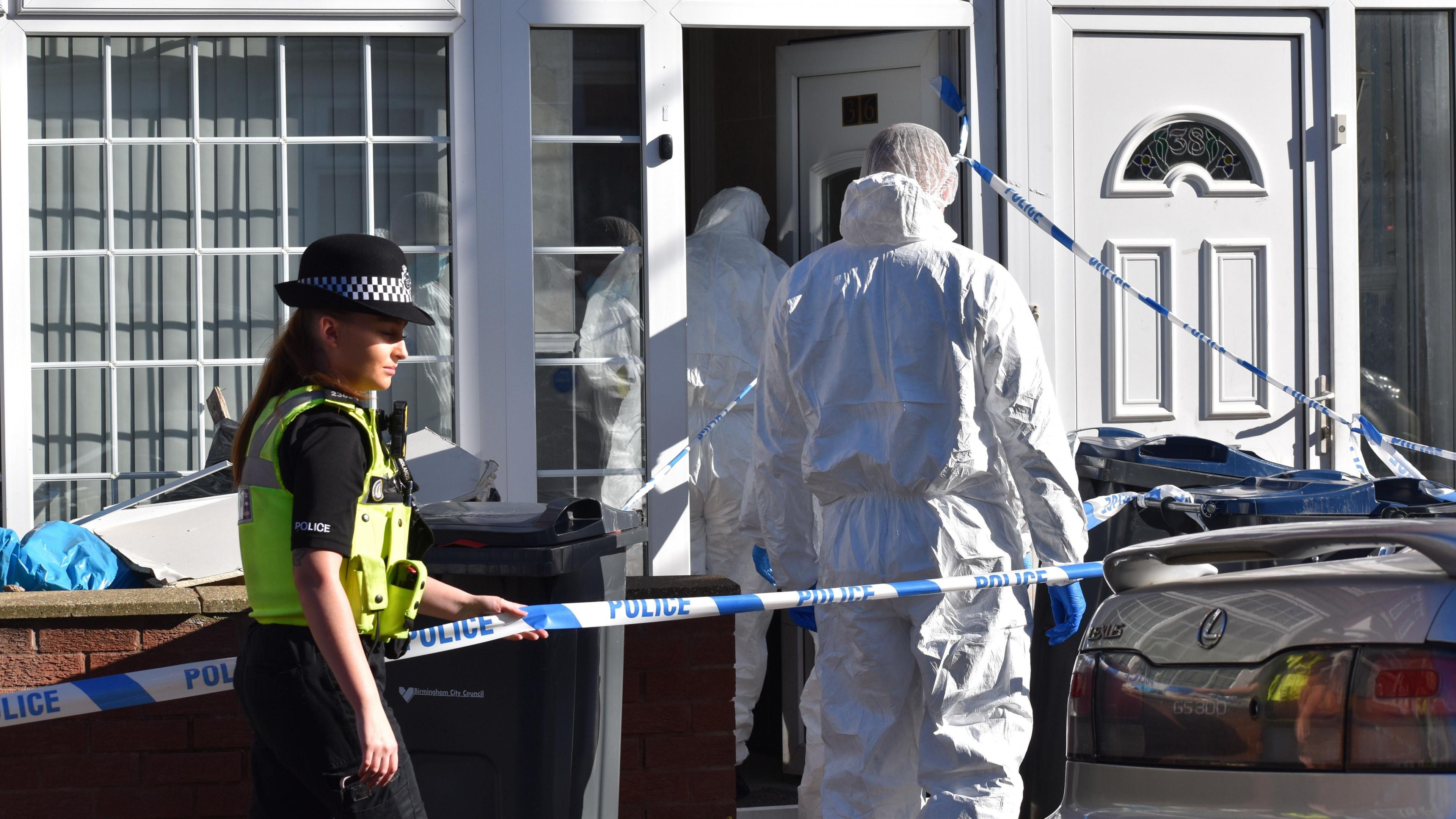 A police cordon around the front of a house. A policewoman with a green vest and black police hat stands by the blue and white police tape with her hand on it. Two forensics officers in all white and wearing blue gloves are standing at the front door of the house.