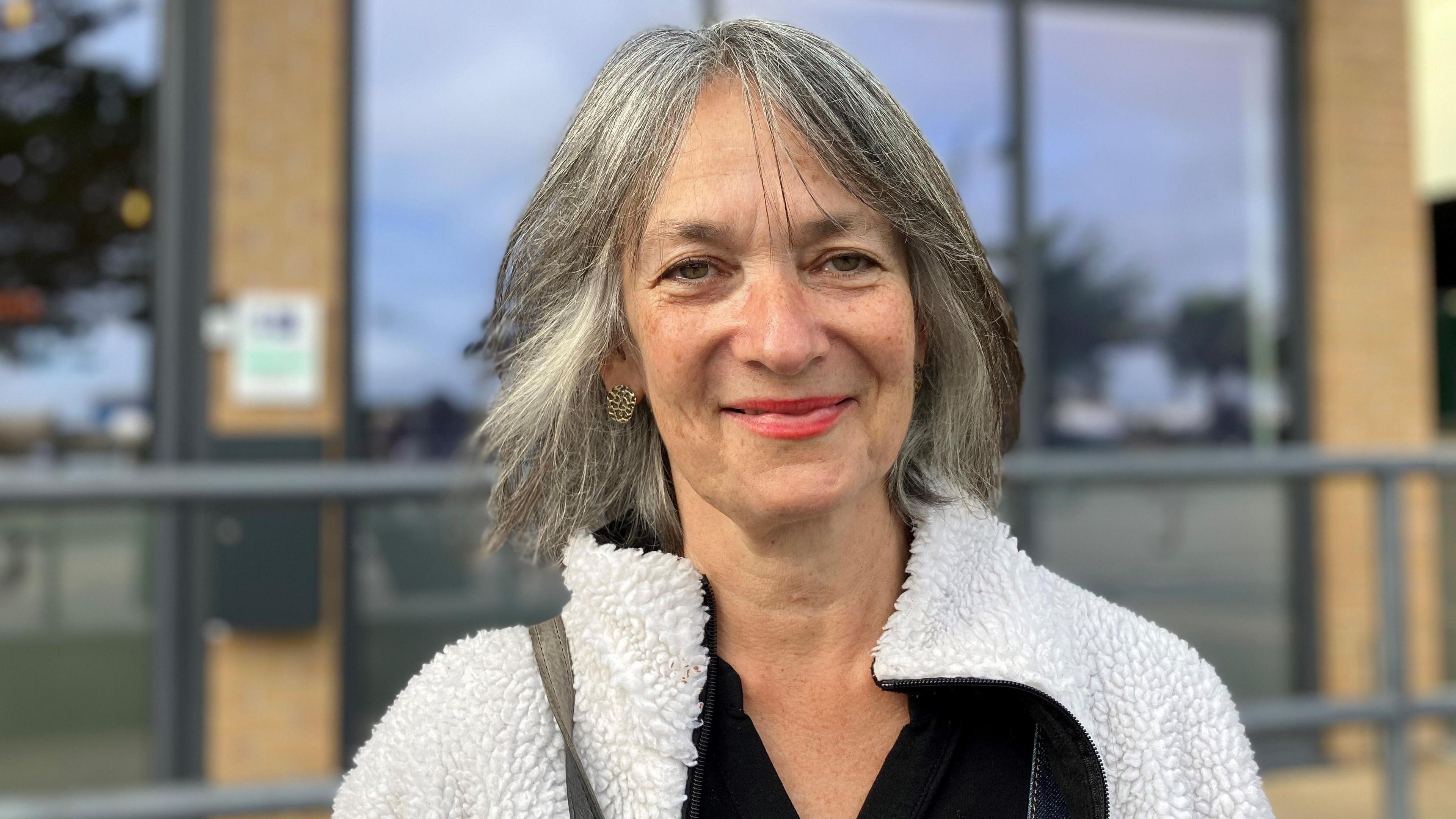 A woman with greying hair in a bob style with brown eyes and wearing red lipstick is smiling at the camera. She has gold earrings and is wearing a white fluffy jacket with a black top underneath.