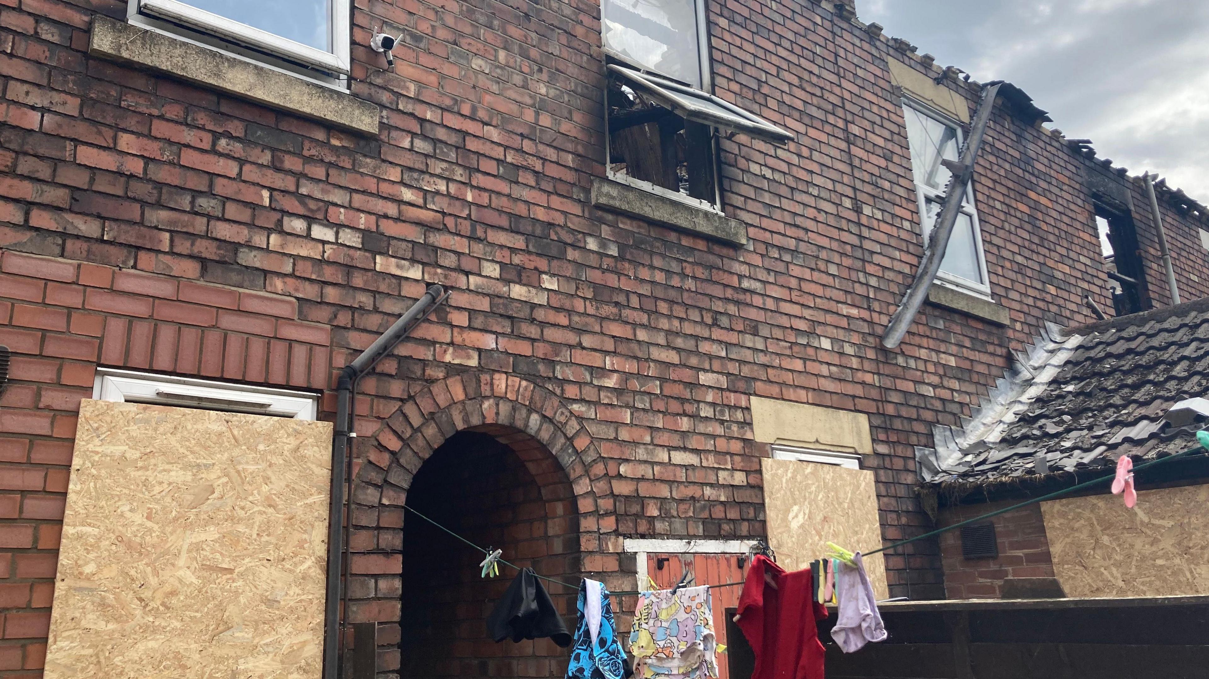 Shot of the rear of a property with boarded up windows and what appears to be smoke damage on the upper floor. There are clothes hung out on a washing line.