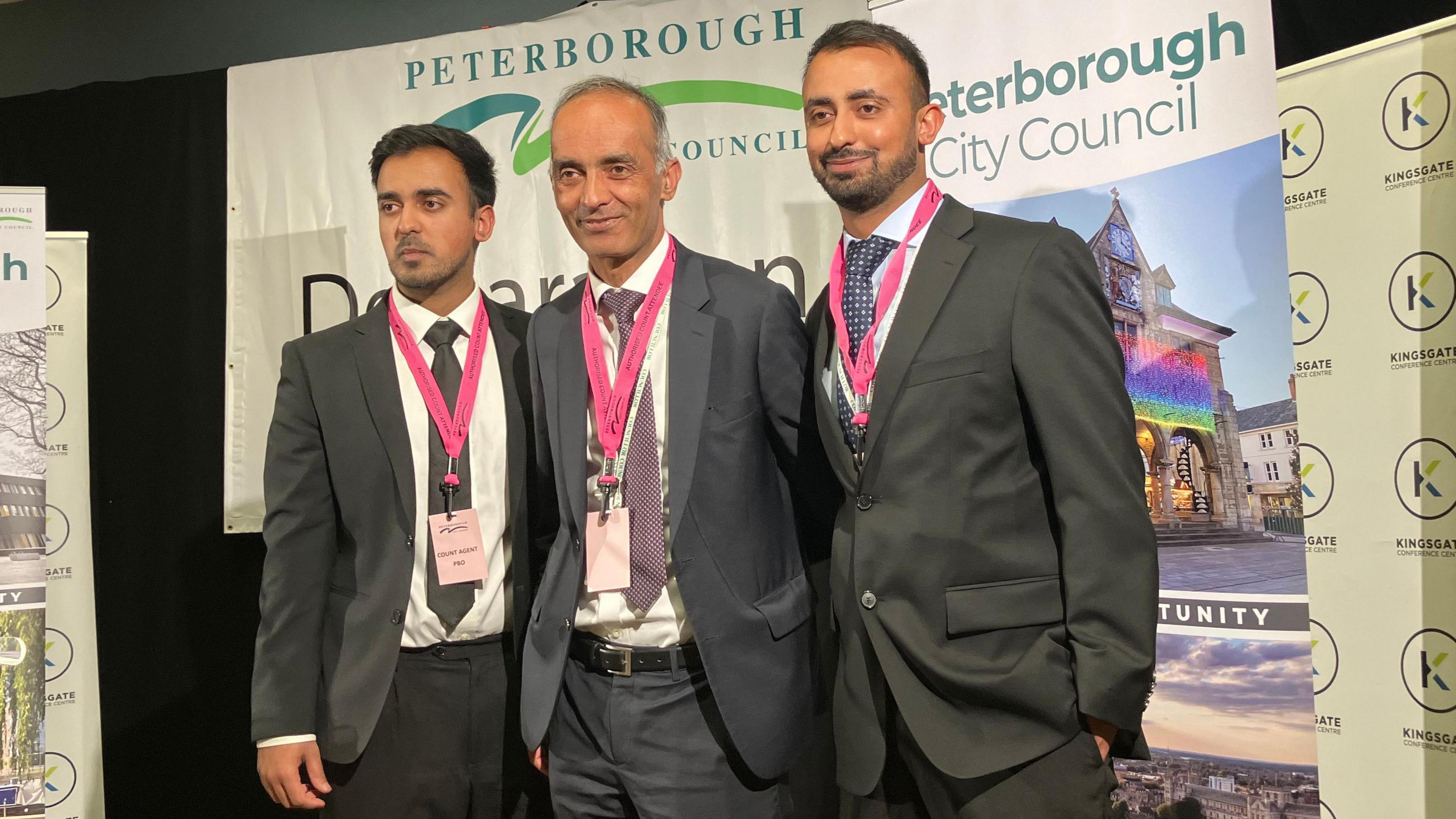 Mohammed Farooq poses for photo alongside two men. They are all dressed in dark suits, with pink lanyard around their necks. They are standing in front of a city council election declaration poster. 