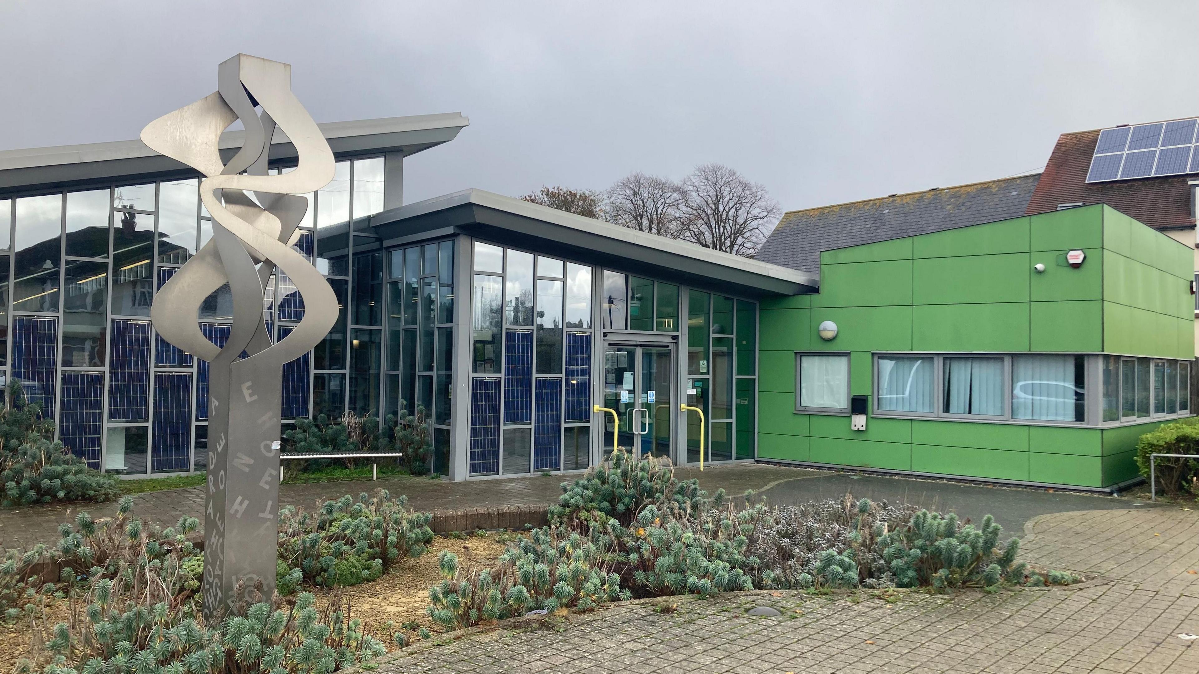 The front of Hamworthy library, Poole. Small shrubs grow beside a metal sculpture 