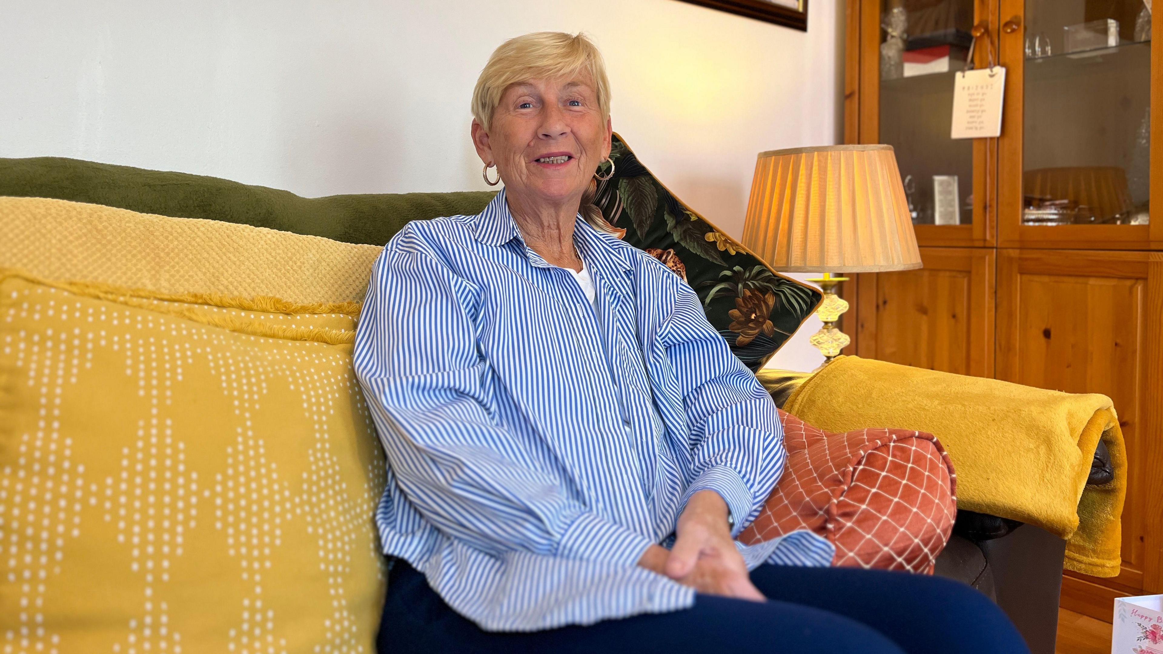 Caron smiles at the camera as she sits on a green sofa with yellow cushions and throws sat next to a lamp