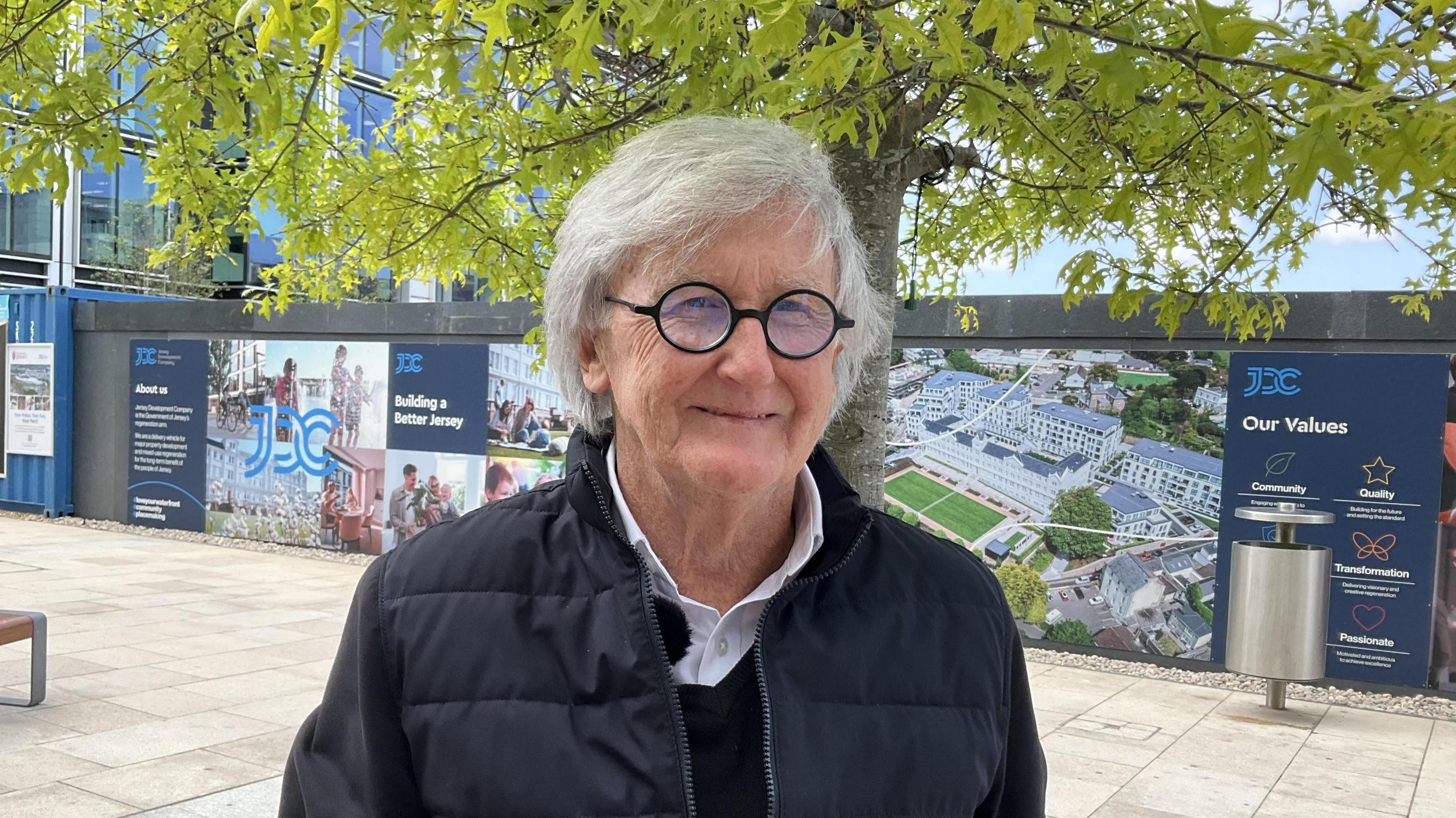 Joe Dickinson pictured smiling at the waterfront business district. He has white hair and round glasses. He's wearing a navy blue jacket and black jumper.