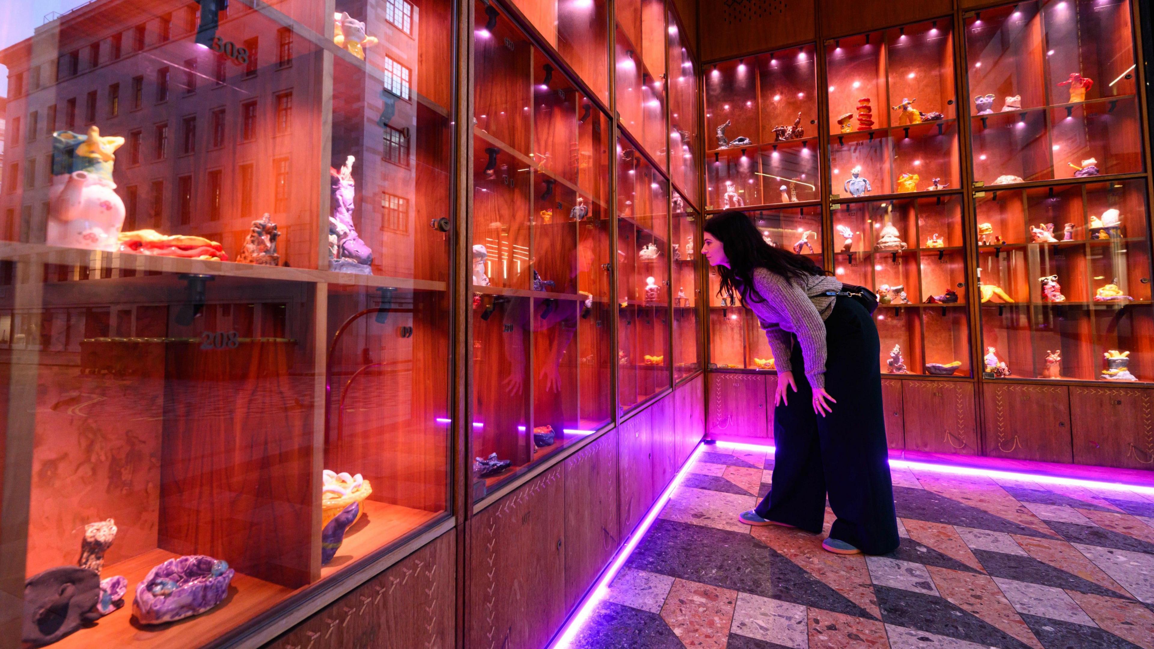 Extraterrestrial-inspired ceramic artworks are on display in wooden cabinets. A woman with long brown hair is looking at them while standing on parquet flooring.