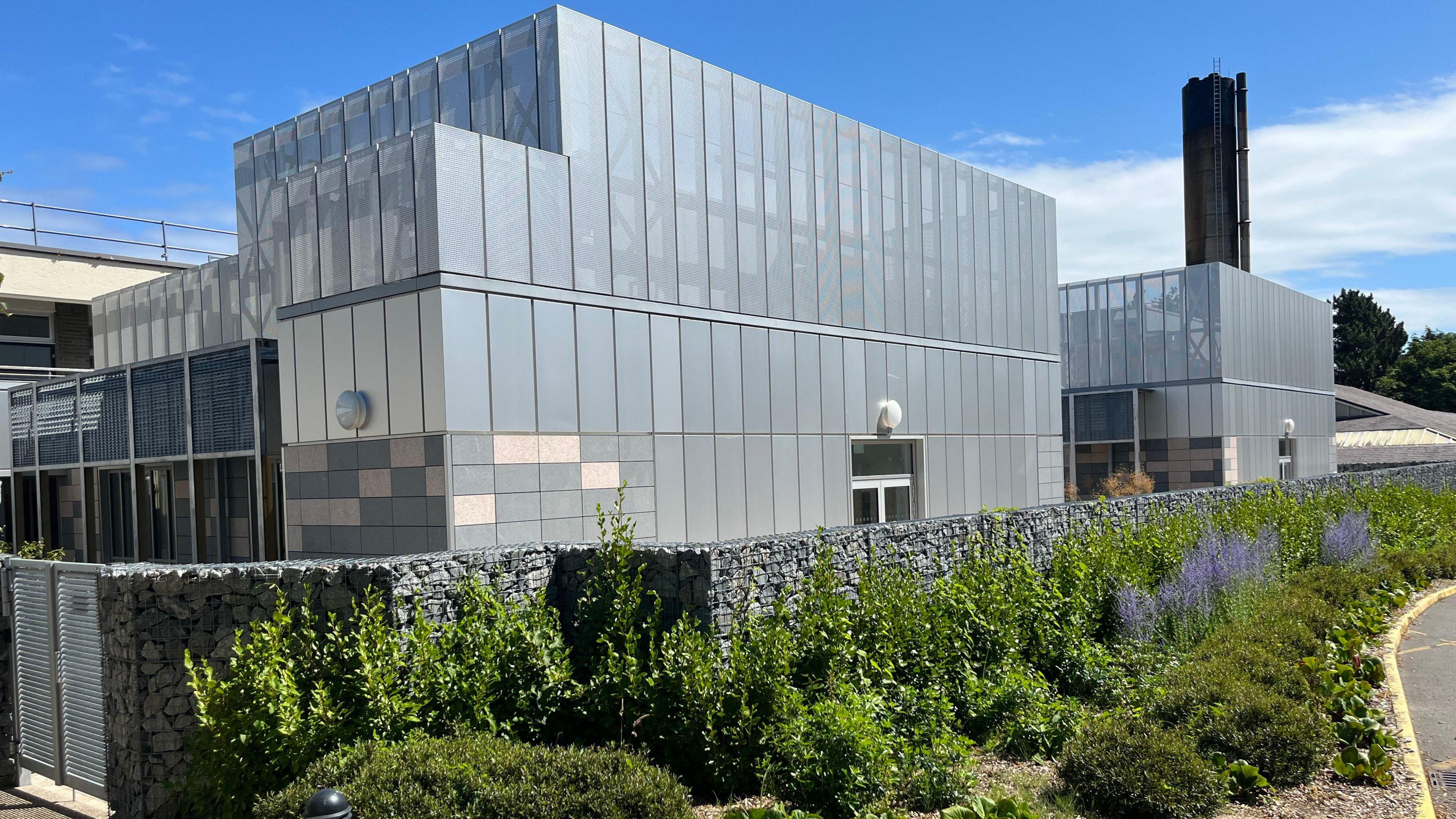 A large grey building, behind some flowers. The Clinical Care Unit at the Princess Elizabeth Hospital. It has a big black chimney rising above it.