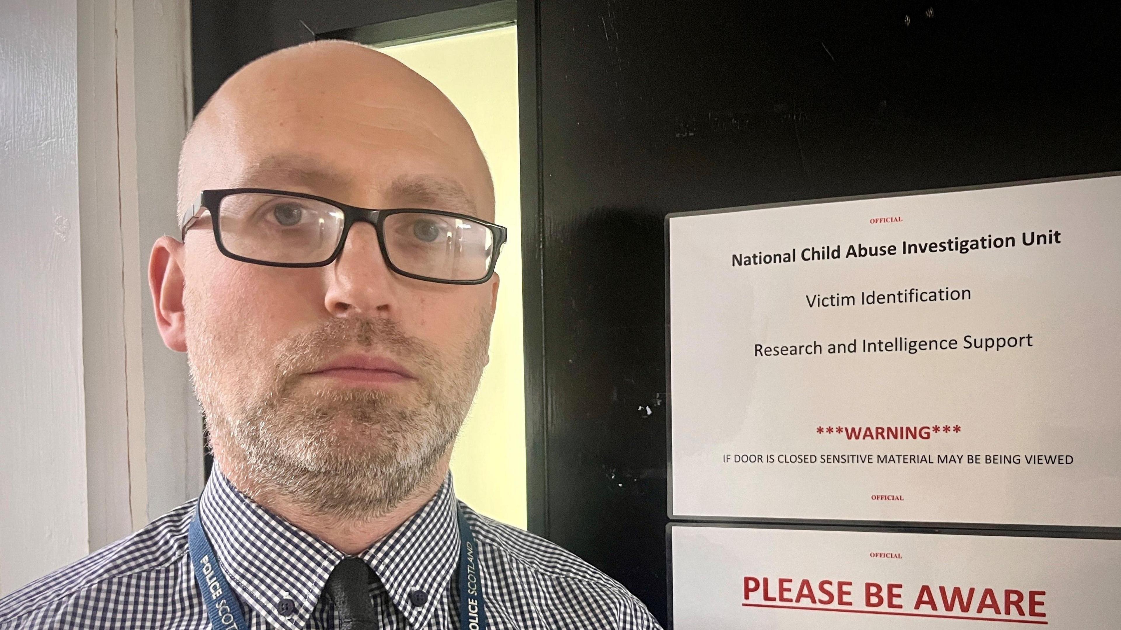 Detective Smith - a bald man with glasses and a beard, looks straight into the camera inside a police station. Behind him on a door, we see a printed sign which reads: "Warning - if door is closed sensitive material may be being viewed."