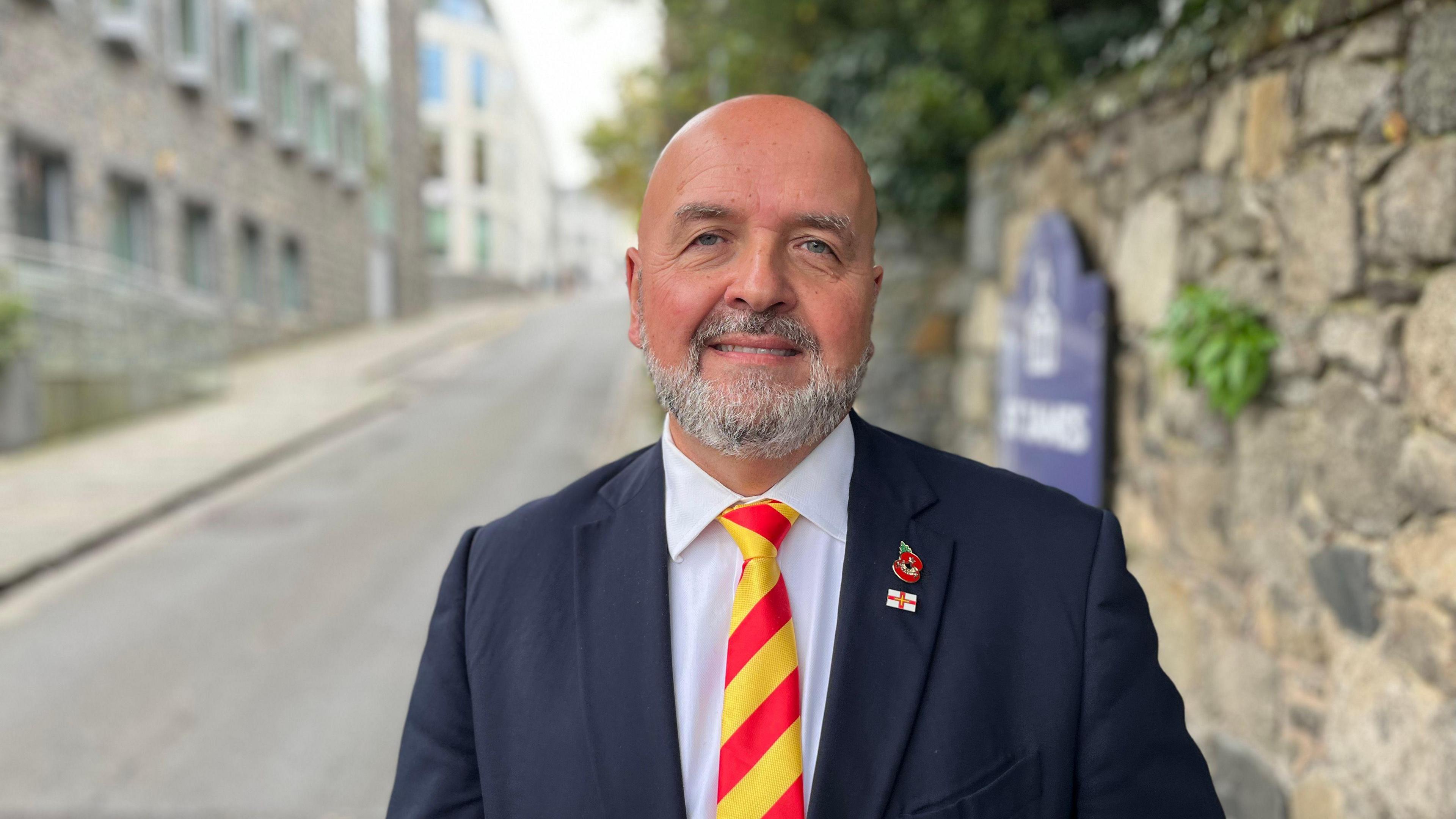 Deputy Jonathan Le Tocq - A white man with a bald head and a grey beard. He's wearing a blue suit, a white shirt, with a red and yellow tie. On the lapel of his suit is a Guernsey pin badge and a poppy. 