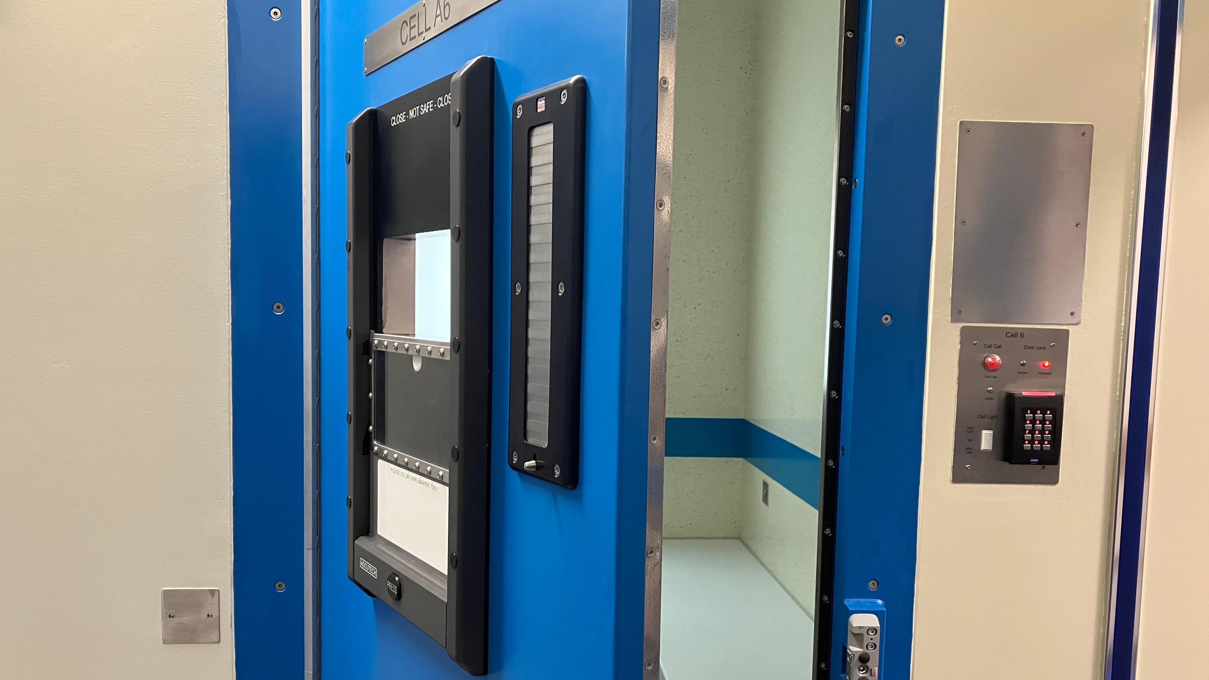 A blue police station cell door that is open with a partial view of inside the cell. A electronic key pad on the righthand side.